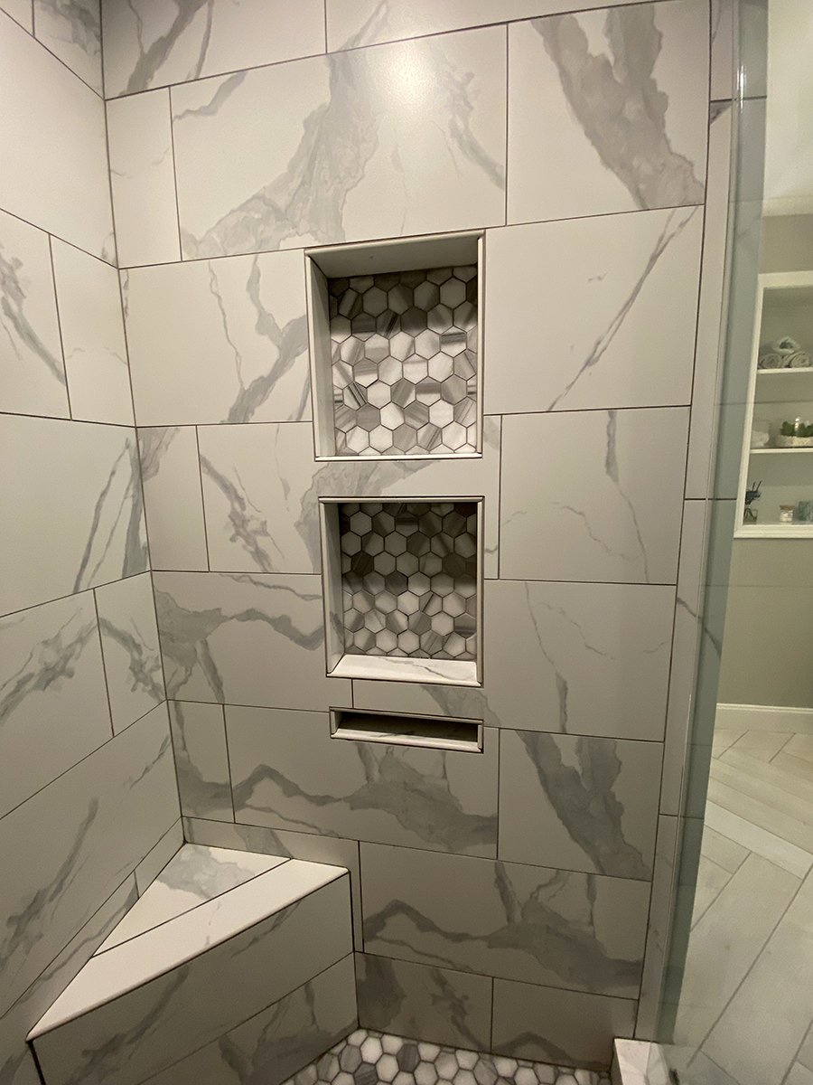 A shower with large white marble tiles with gray veining, two recessed shelves with hexagonal tile backs, and a built-in bench in the corner.