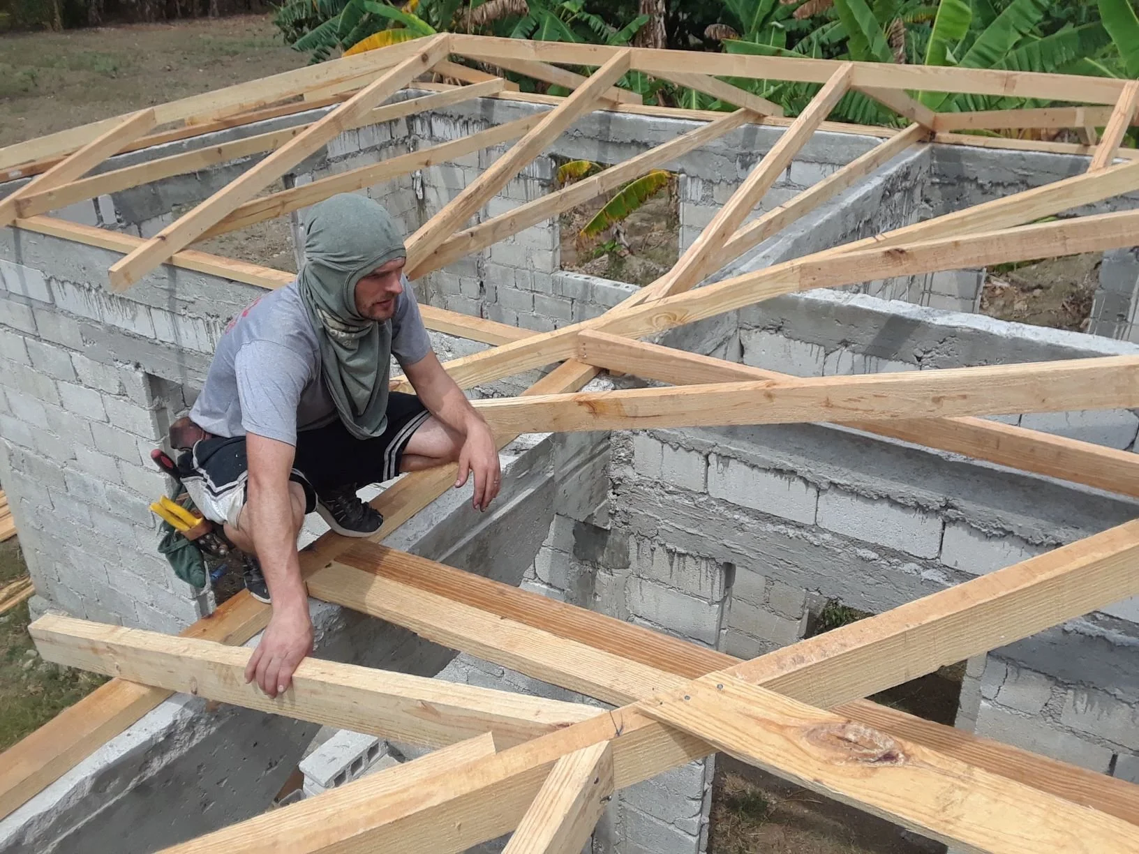 A man with a gray bandana, gray T-shirt, and black shorts crouches on a wooden beam atop a partially constructed brick wall, working on the roof framework with wooden rafters.