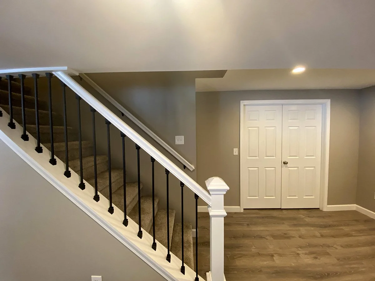 Interior view of a home with stairs leading up, a hallway with a wooden floor, and a white double door in a beige wall.