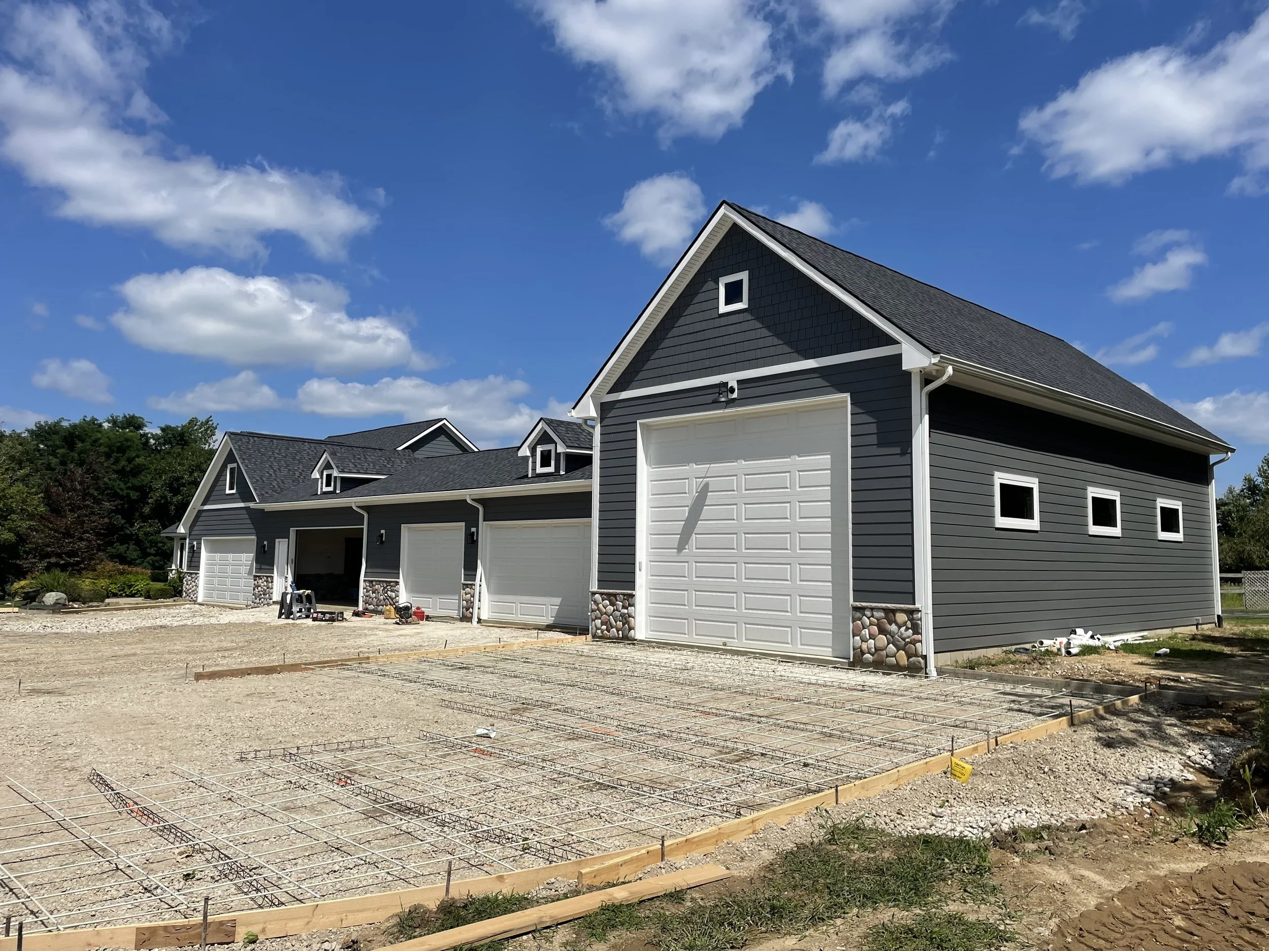 New gray house with white trim under construction, with a concrete foundation, small windows, and a blue sky with clouds.