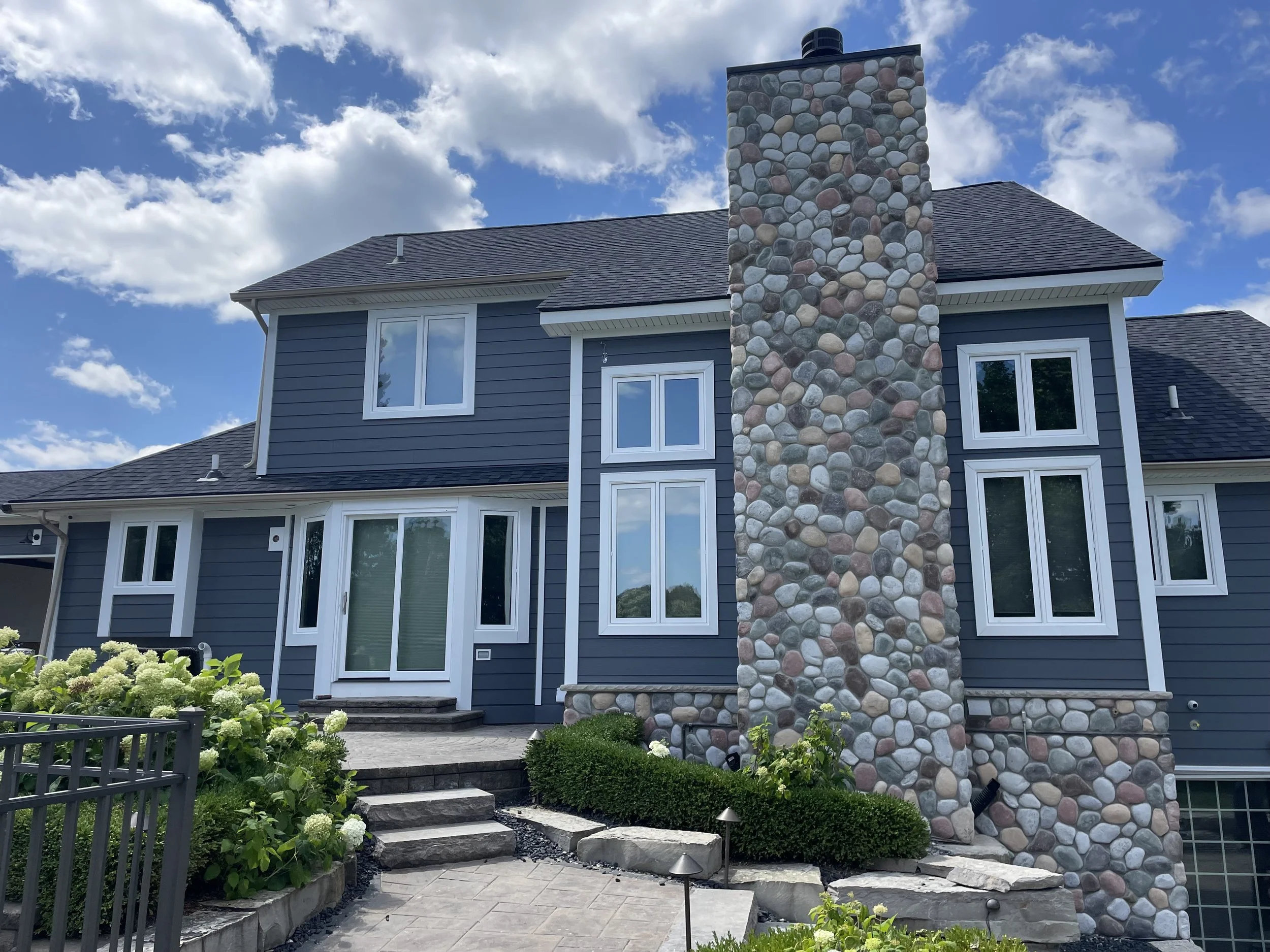 Front view of a blue, two-story house with a stone chimney, multiple windows, a sliding glass door, and a landscaped yard with bushes, flowers, and a stone pathway under a partly cloudy sky.