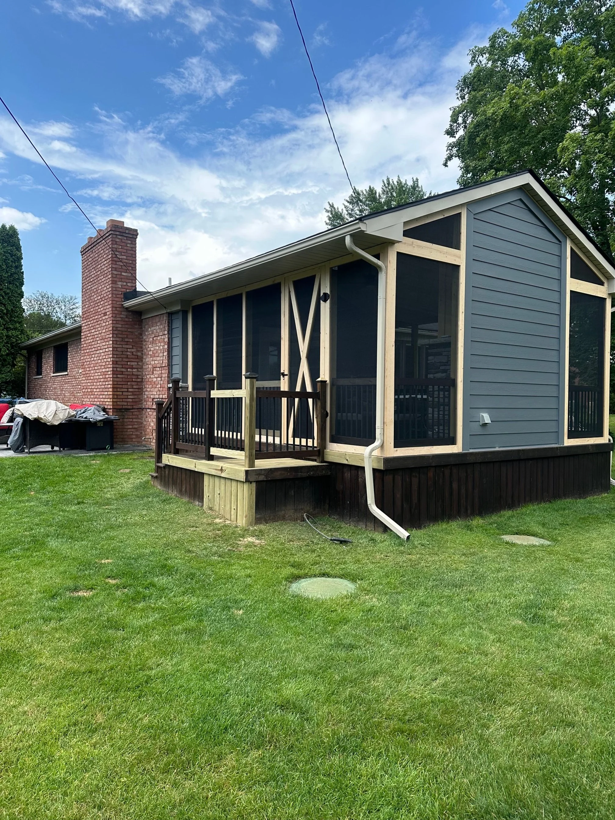 A house with a brick chimney, a porch with a wooden railing, and screened-in area under construction. Green lawn and trees in the background, blue sky with some clouds.