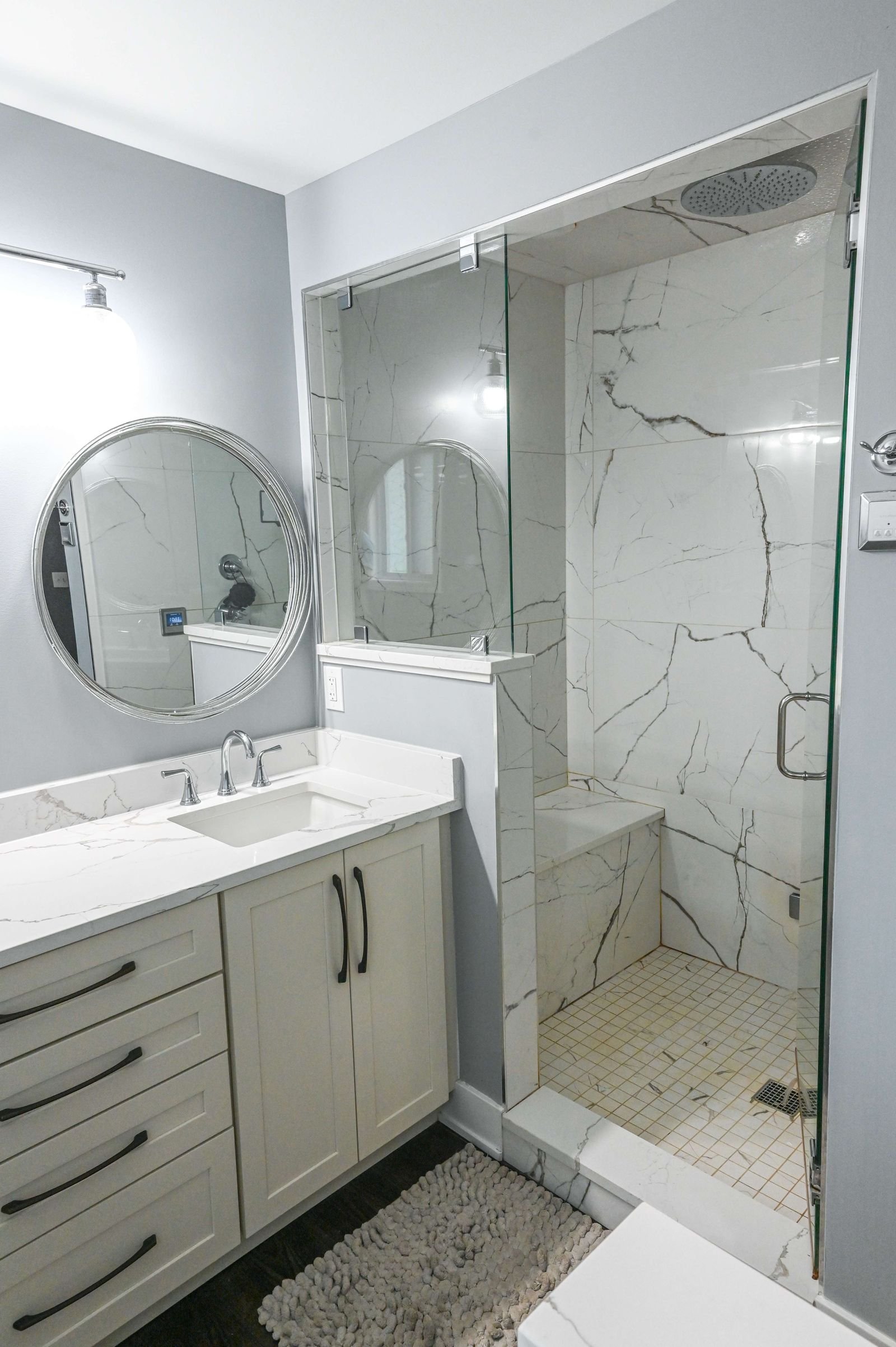 Modern bathroom with a vanity, round mirror, marble countertop, and a glass-enclosed shower with marble tiles and a built-in bench.