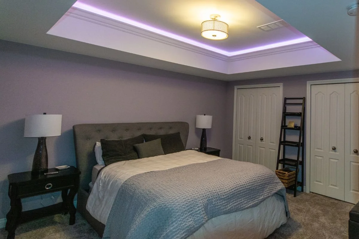 A bedroom with a vaulted ceiling featuring purple LED lighting and a central ceiling light fixture. It has a bed with a tufted headboard, black and gray pillows, beige bedding, and two black nightstands with lamps. There are double closet doors and a