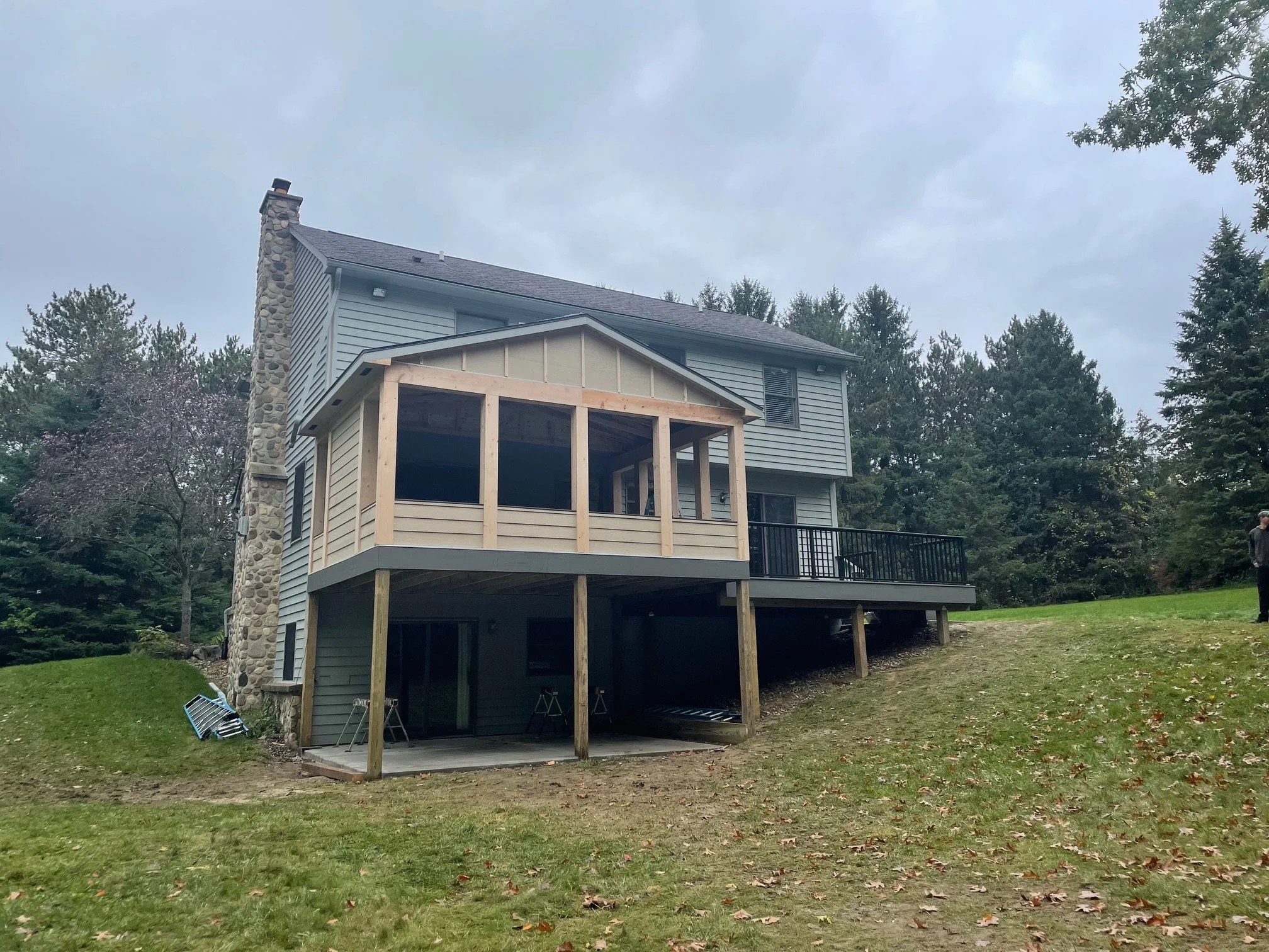 House with a partially built screened-in porch extension on a grassy yard, surrounded by trees and cloudy sky.