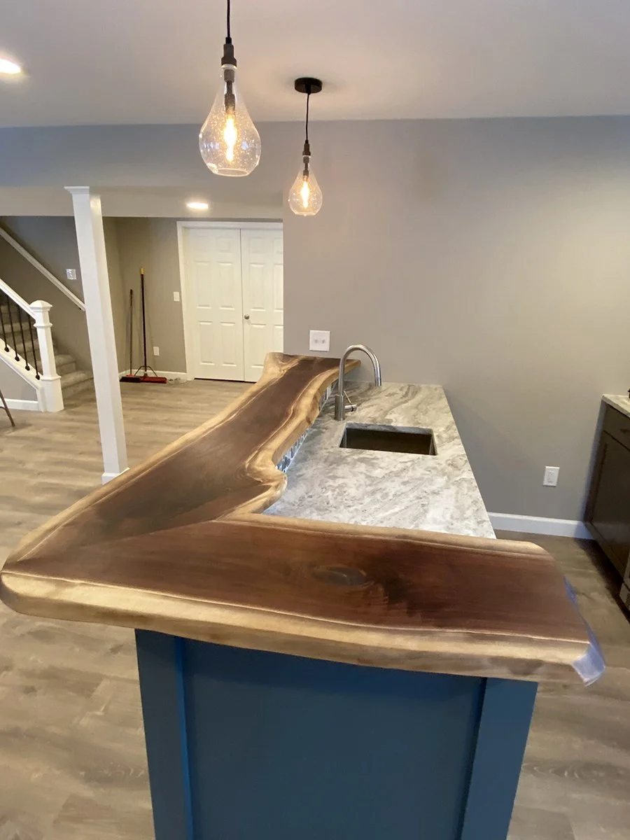 Kitchen island with a natural wood countertop, modern gray cabinetry, and a marble or granite countertop with a sink. Pendant lights hang above the island, and a staircase is visible in the background.
