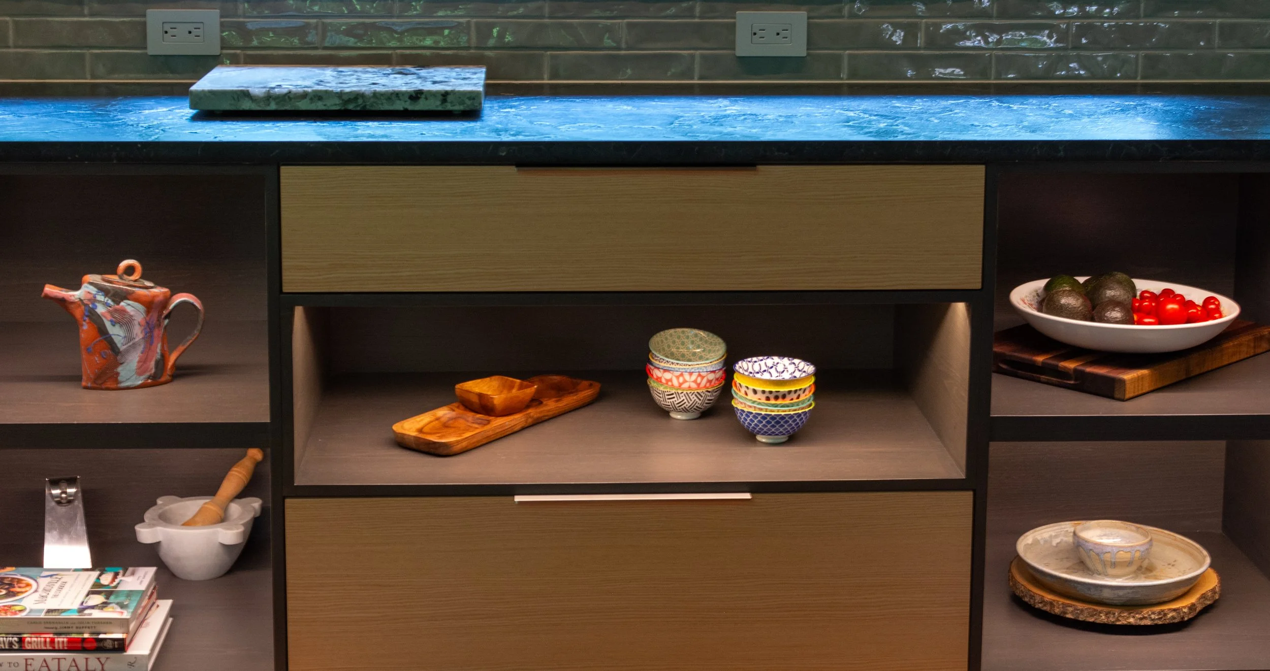 Interior of a modern kitchen cabinet with kitchenware, bowls, teapot, and decorative items. A teal countertop with electrical outlets above.