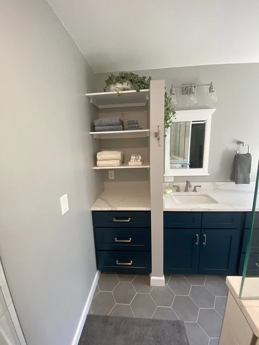 Bathroom with dark blue vanity, white marble countertop, open shelves with folded towels and greenery, mirror, modern light fixture, and hexagonal gray floor tiles.