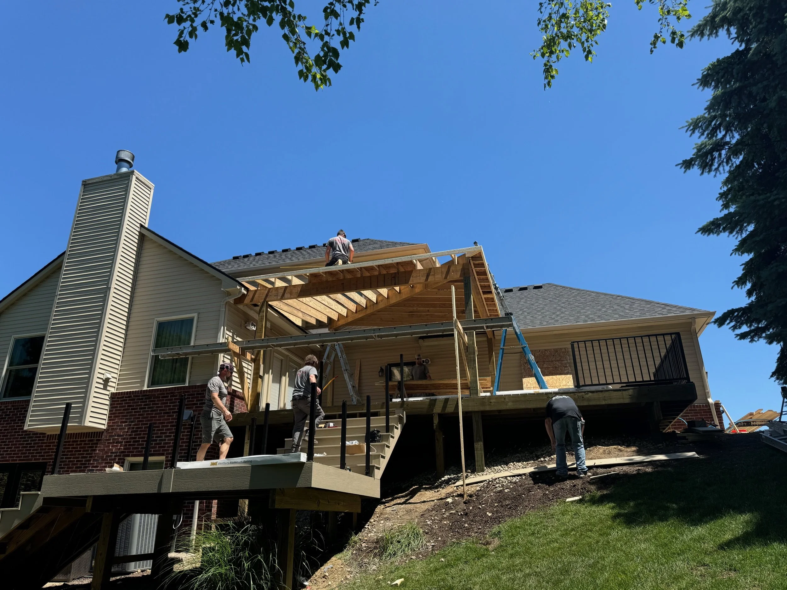 Construction workers building a deck extension on a two-story house under a clear blue sky.