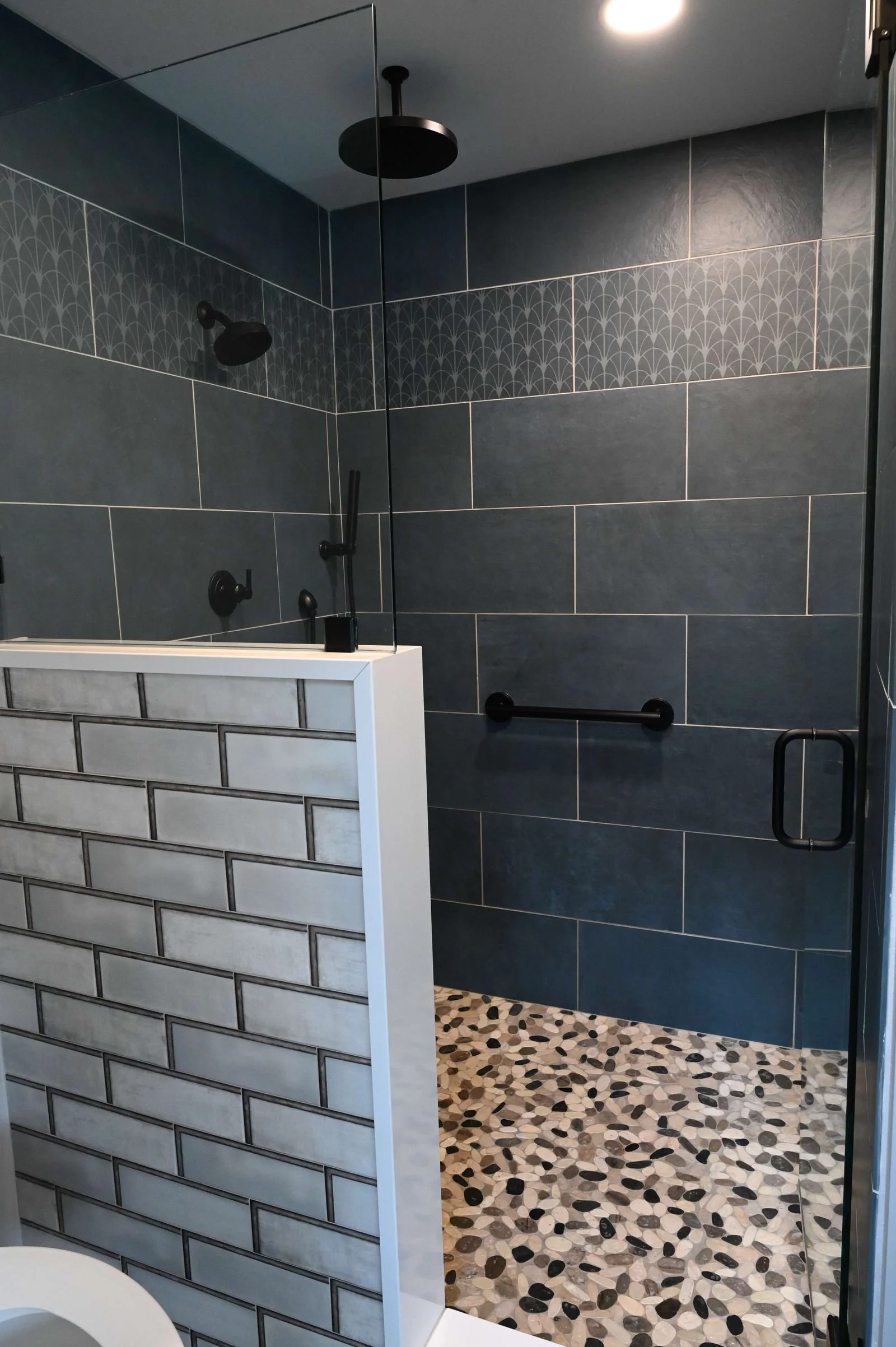 Modern bathroom with a walk-in shower featuring dark gray tiles, black fixtures, a pebble patterned floor, and a half wall with subway tiles.