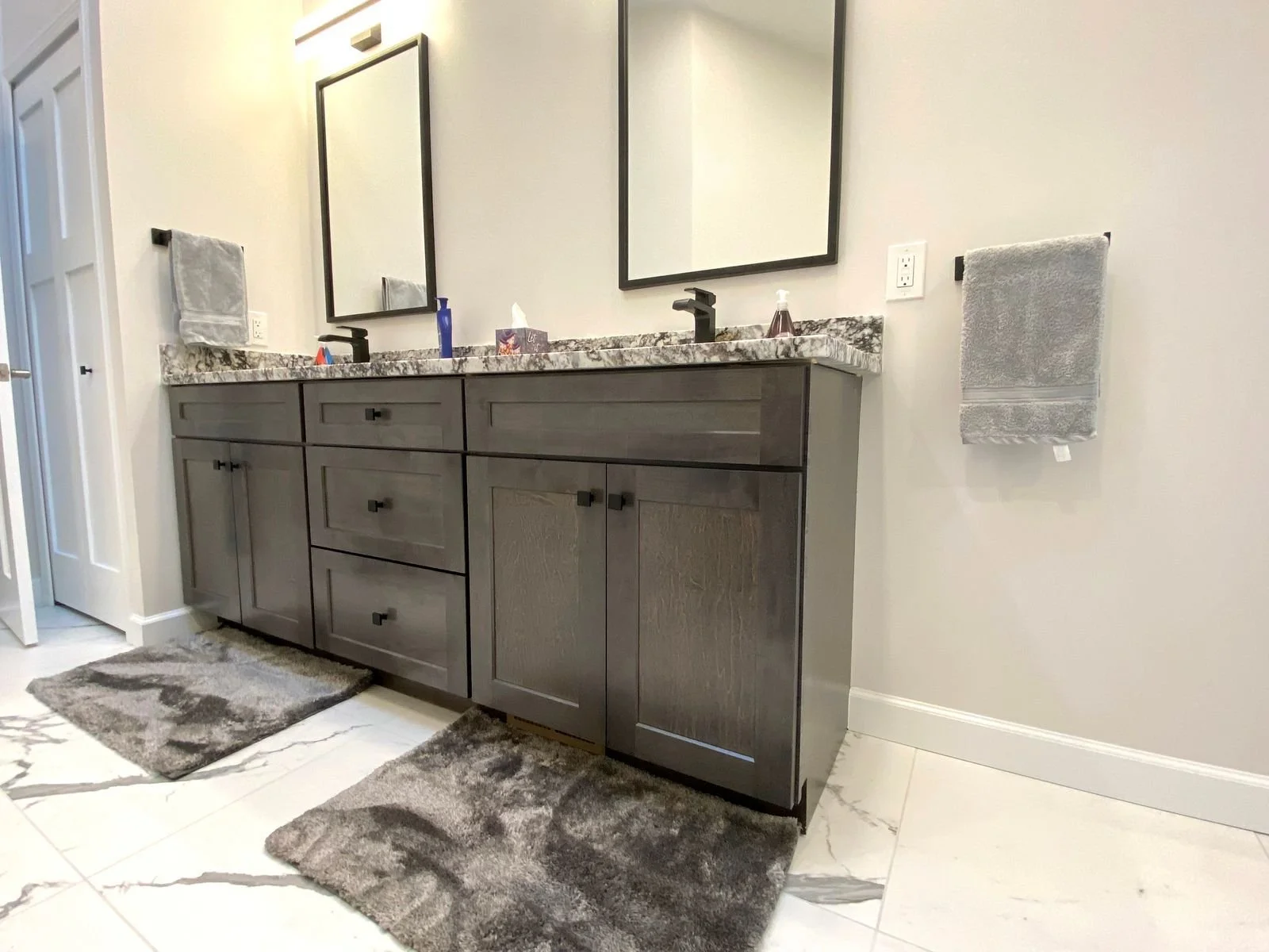 Bathroom sink vanity with two mirrors, granite countertop, gray cabinets, towels on either side, and bathroom accessories on the counter.