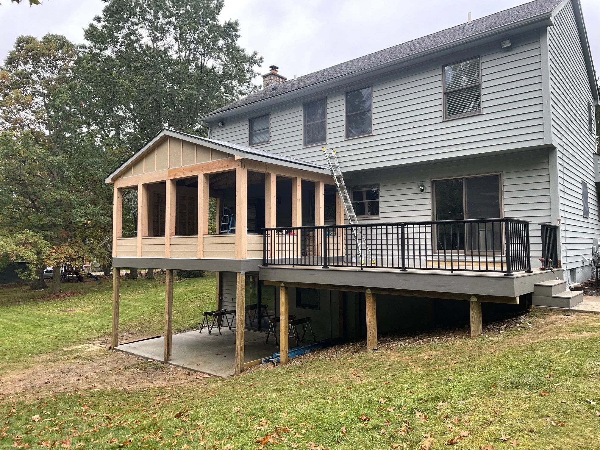 A two-story house with an expanded deck in progress, featuring a screened-in porch under construction, a ladder leaning against the house, and tools on the deck, with a tree and grassy yard in the background.