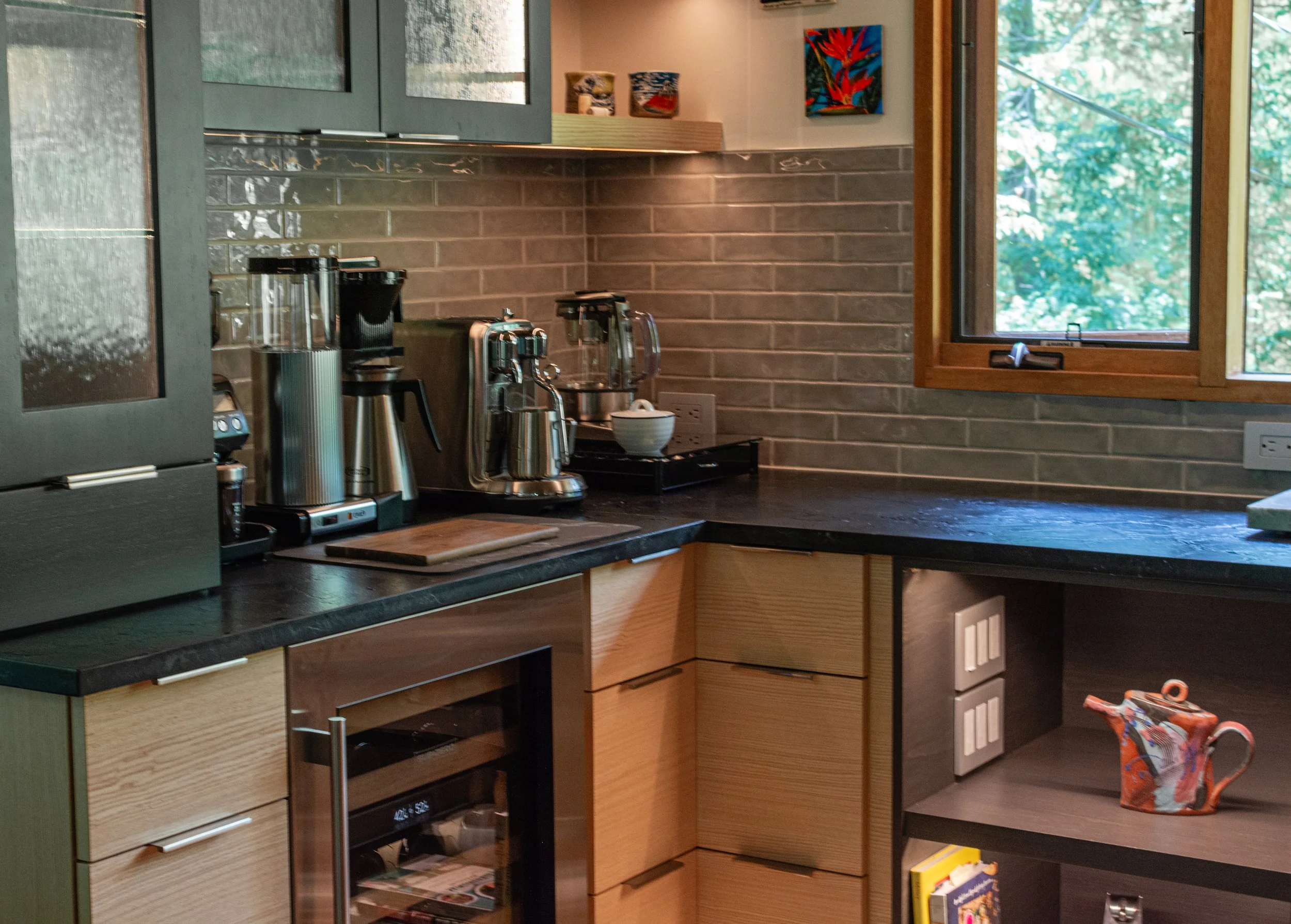 Kitchen with black countertop, gray brick backsplash, coffee makers, and an open window showing green trees outside.