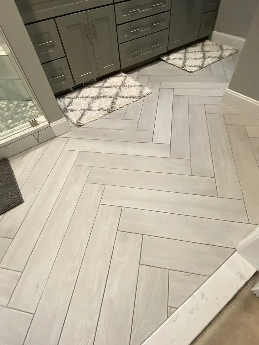 Living room with light-colored herringbone wood flooring, a gray cabinet, two patterned rugs, and a glass shower door.