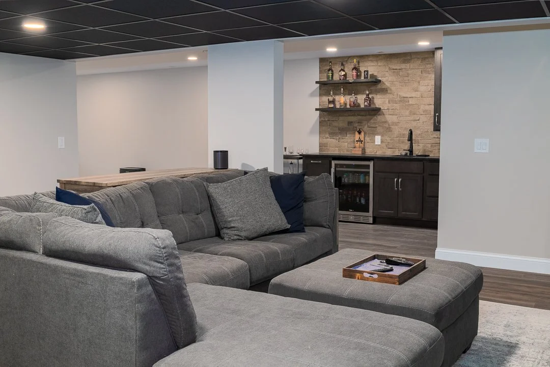 Living room with a large gray sectional sofa and an ottoman. In the background, a bar area with shelves, liquor bottles, and a mini fridge, with a brick wall and a black countertop.