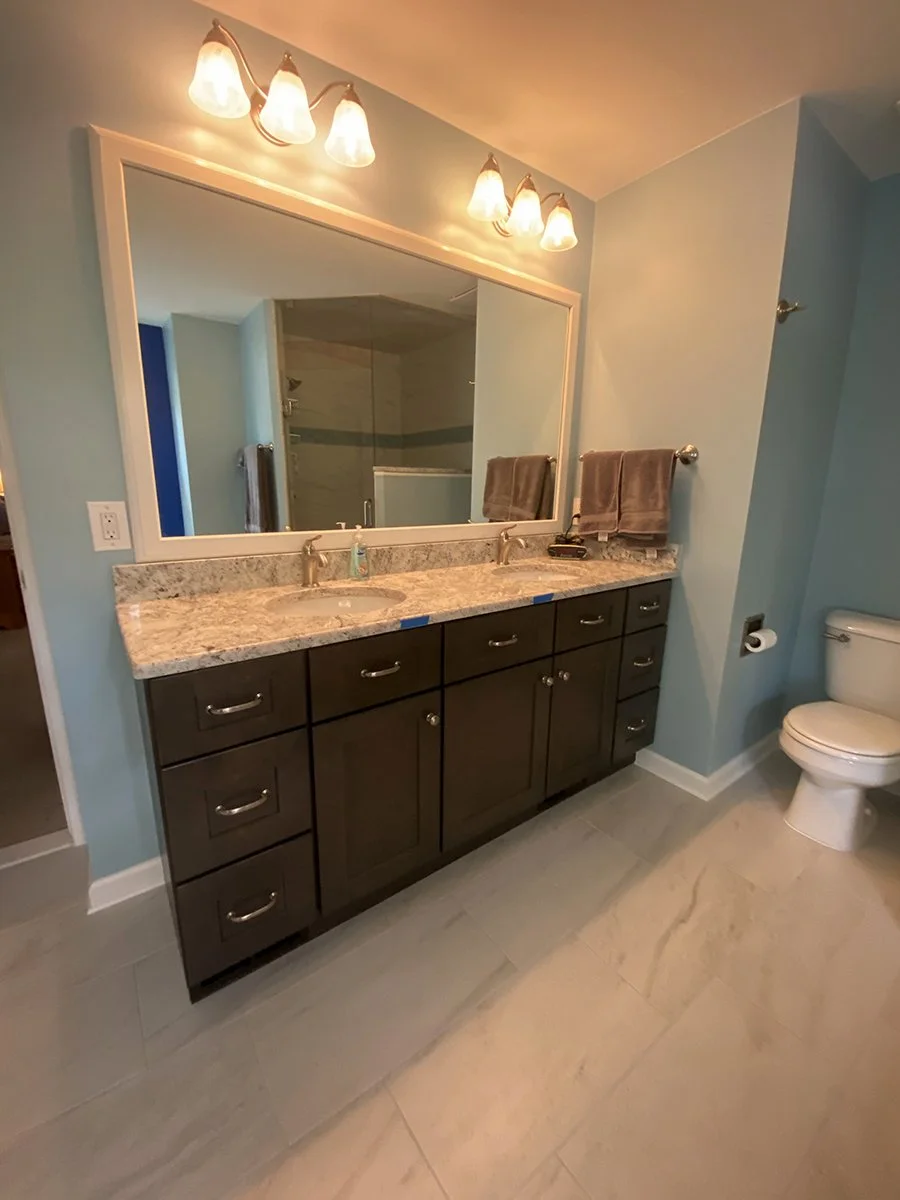 Bathroom with a double vanity mirror, granite countertop, and dark wood cabinets. There are two sinks with gold fixtures, two towel bars with brown towels, and a large mirror above the vanity. To the right is a white toilet and a toilet paper holder.