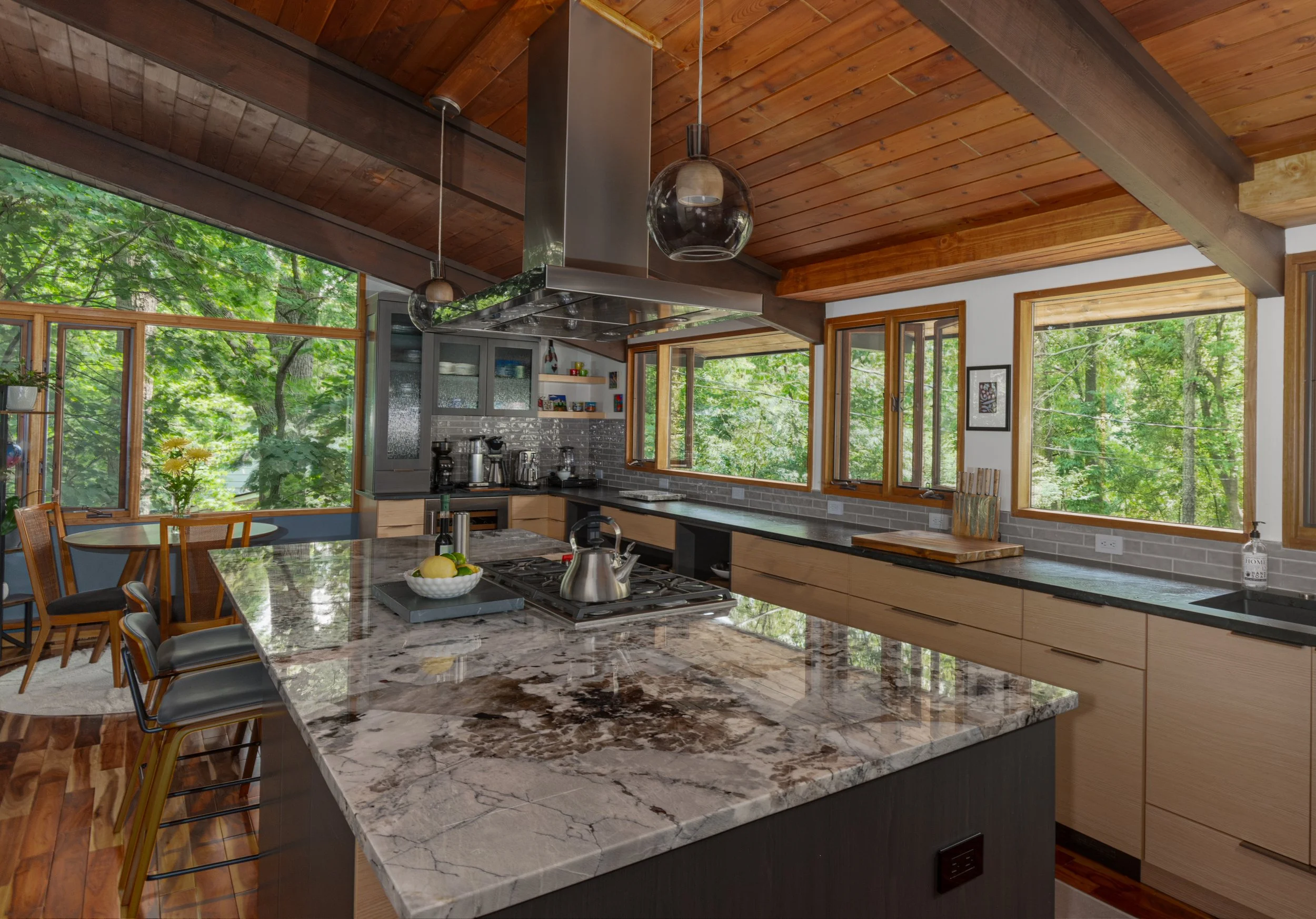 Modern kitchen with large central island with marble countertop, situated under a wooden ceiling with large windows overlooking a green, wooded landscape.