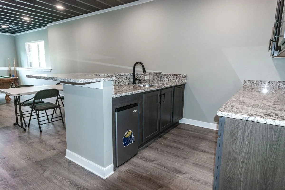 Kitchen area with granite countertops, dark cabinets, small refrigerator with a sticker, and a sink with a black faucet. In the background, there is a table with two chairs and a pool table near the window.