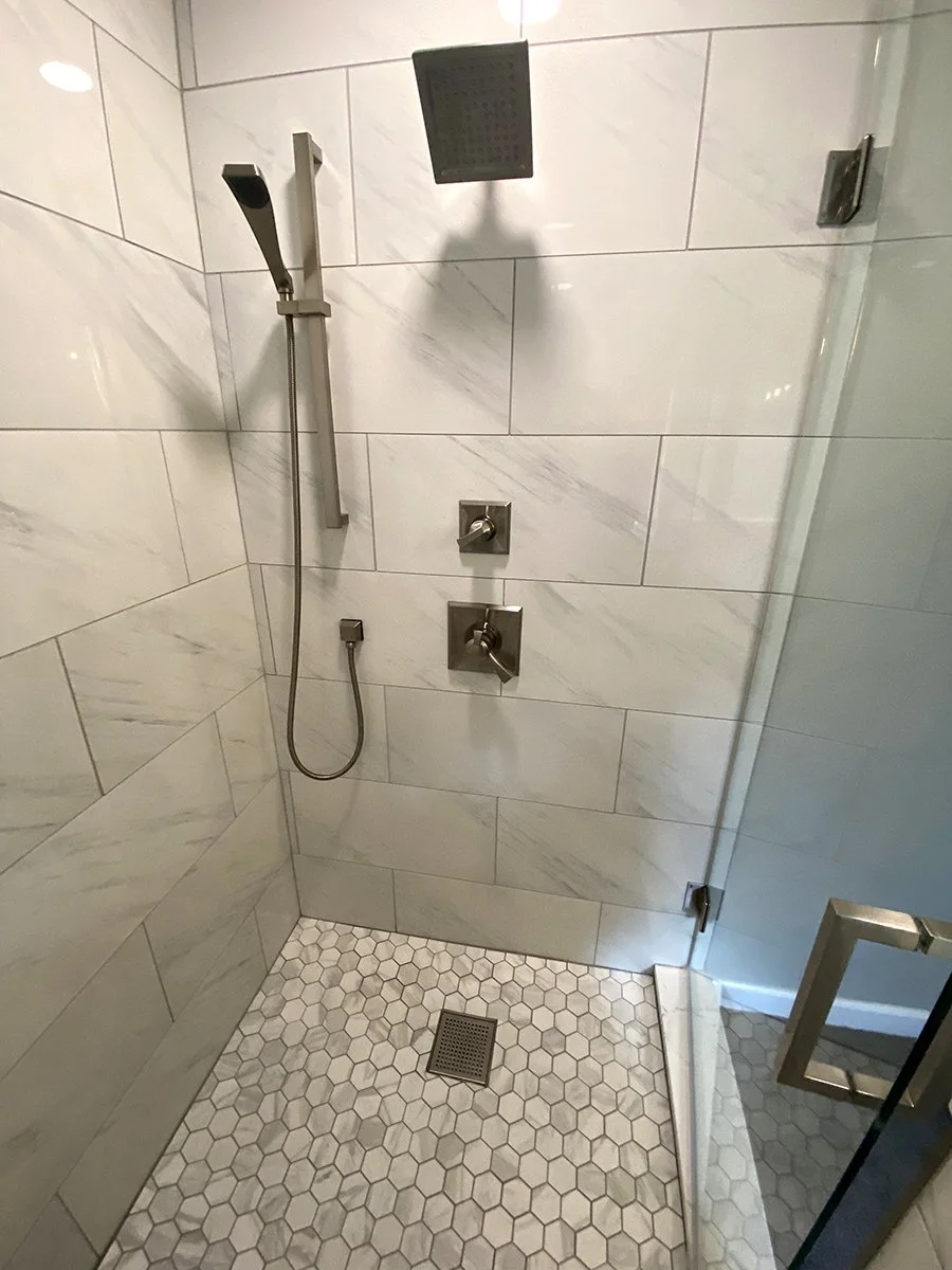 Shower with white marble tiles, a square rain shower head, a handheld showerhead, controls, and a hexagon tile floor with a drain.
