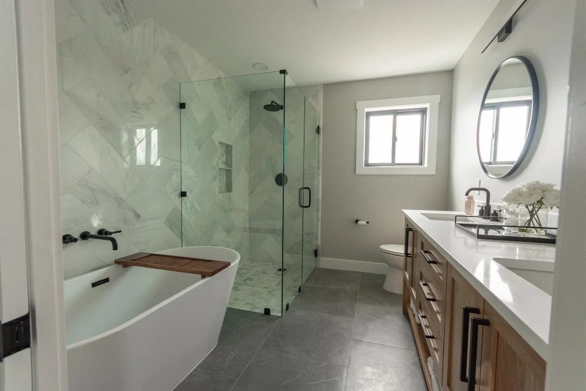 Modern bathroom with a bathtub, a glass-enclosed shower, a toilet, and a wooden vanity with a sink and round mirror. The room has light gray walls, a window, and a gray tiled floor.