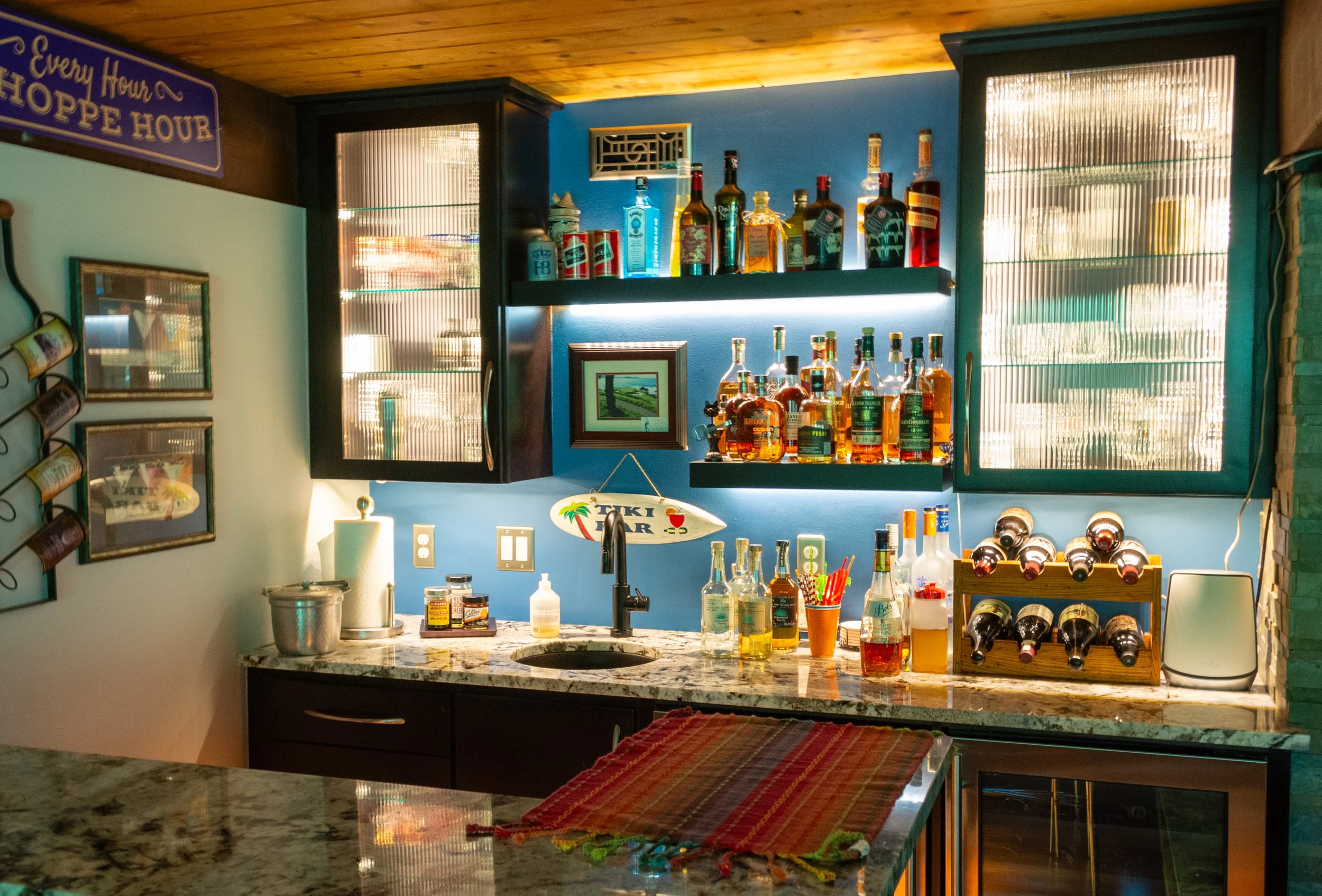 Home bar with bottles of liquor on shelves, wine bottles in a rack, and bar tools on a marble countertop, decorated with tropical-themed items and framed pictures.