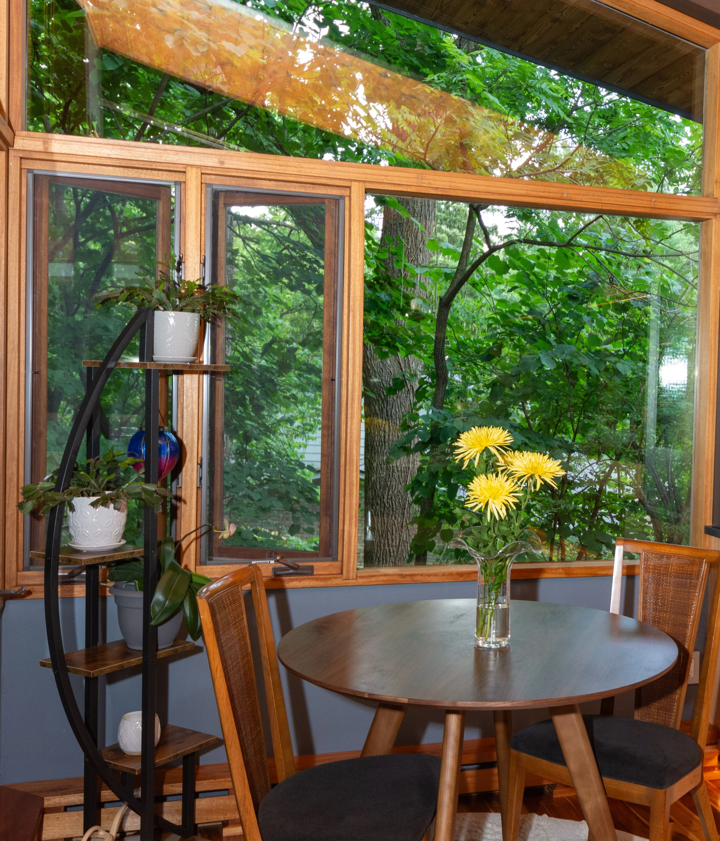A dining area with a round wooden table, a vase of yellow flowers, and wooden chairs, next to a large open window showing green trees outside.
