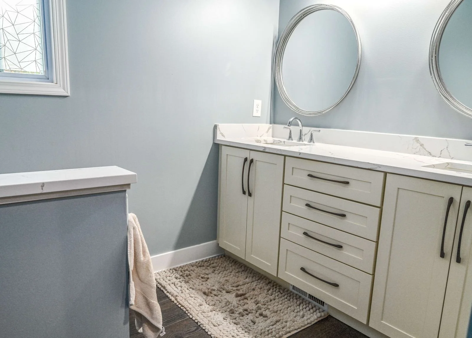 A bathroom vanity with a white marble countertop, a sink with a silver faucet, and two oval mirrors above. Light blue walls, a window with frosted glass, and a beige textured rug on dark wood floor. A towel hangs on the side of a small blue cabinet.