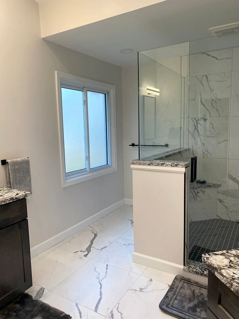 Modern bathroom with white marble floor tiles, a glass-enclosed shower with marble walls, a window with frosted glass, and dark wood cabinets with granite countertops.