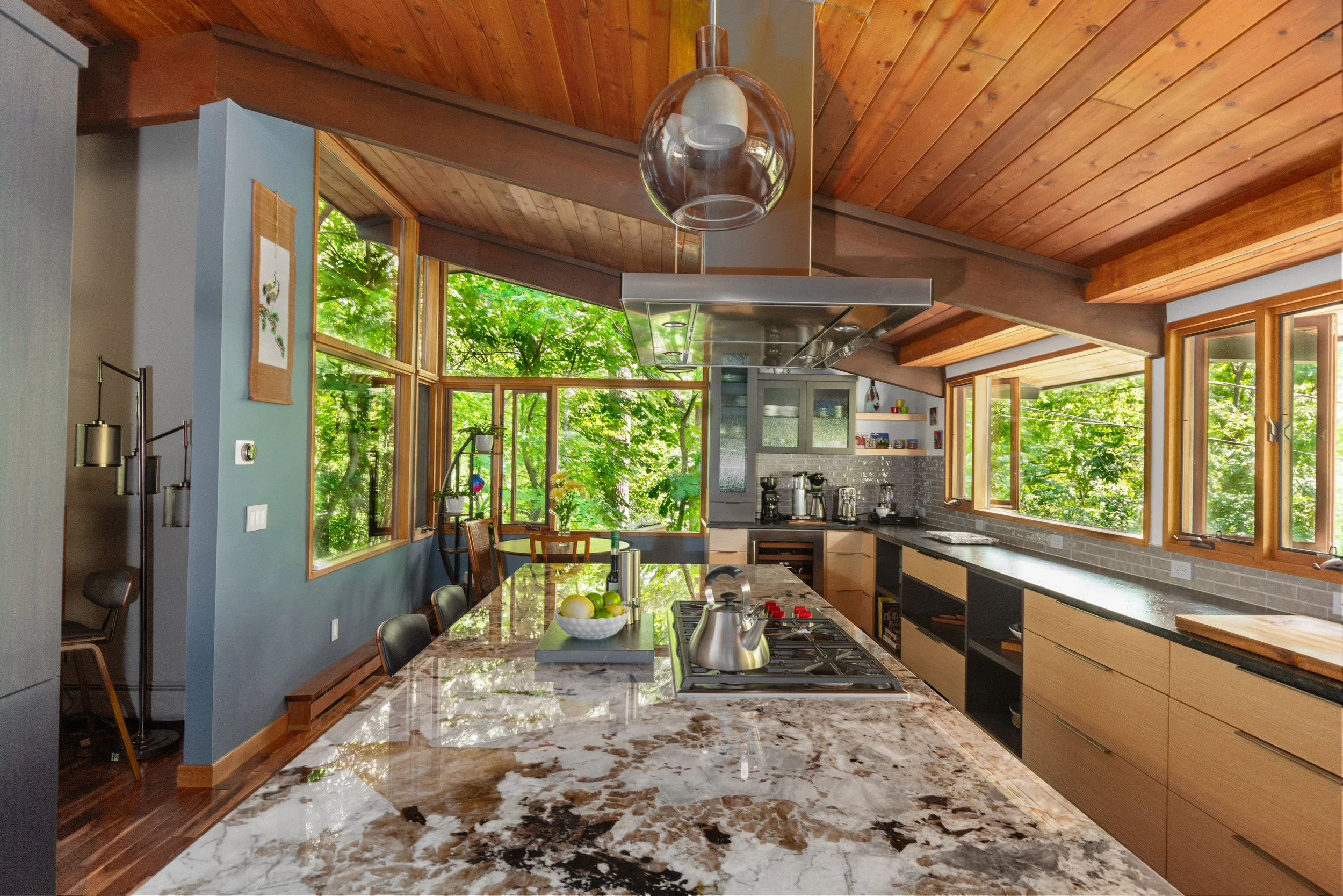 Modern kitchen with marble countertop, wooden cabinets, large windows, and green outdoor scenery.