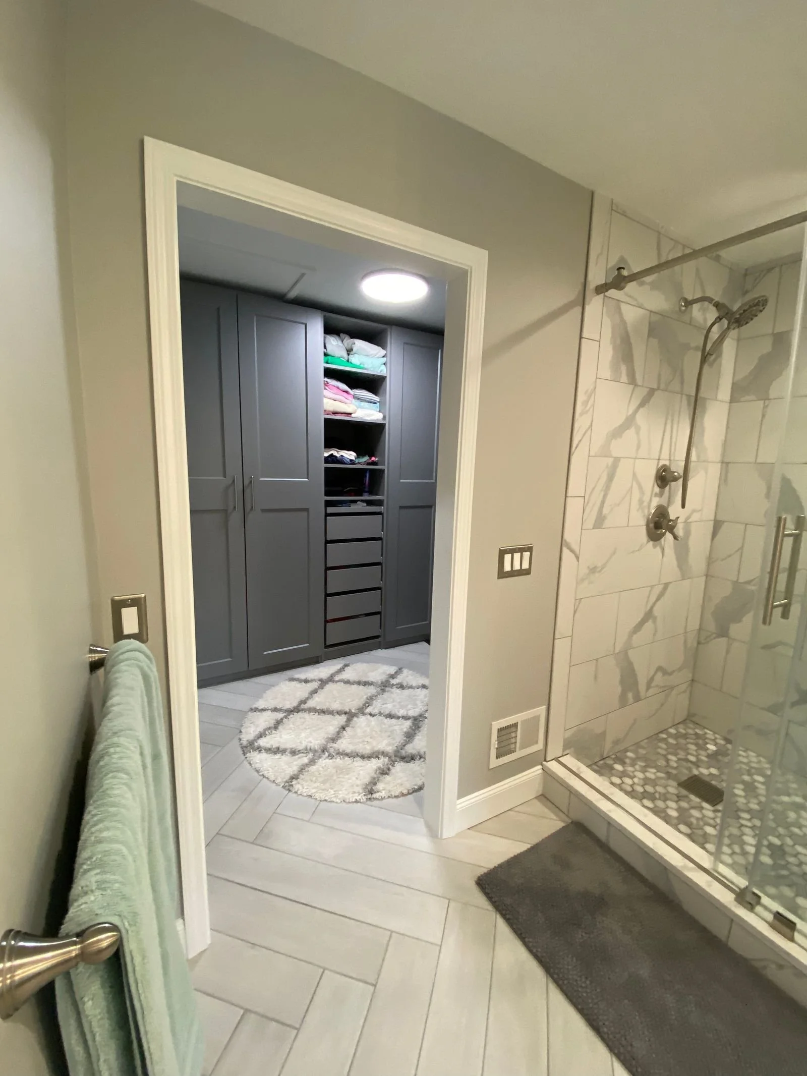 Bathroom with a shower stall featuring marble tiles, a gray towel hanging on a towel bar, and a walk-in closet with gray cabinets and a round rug.