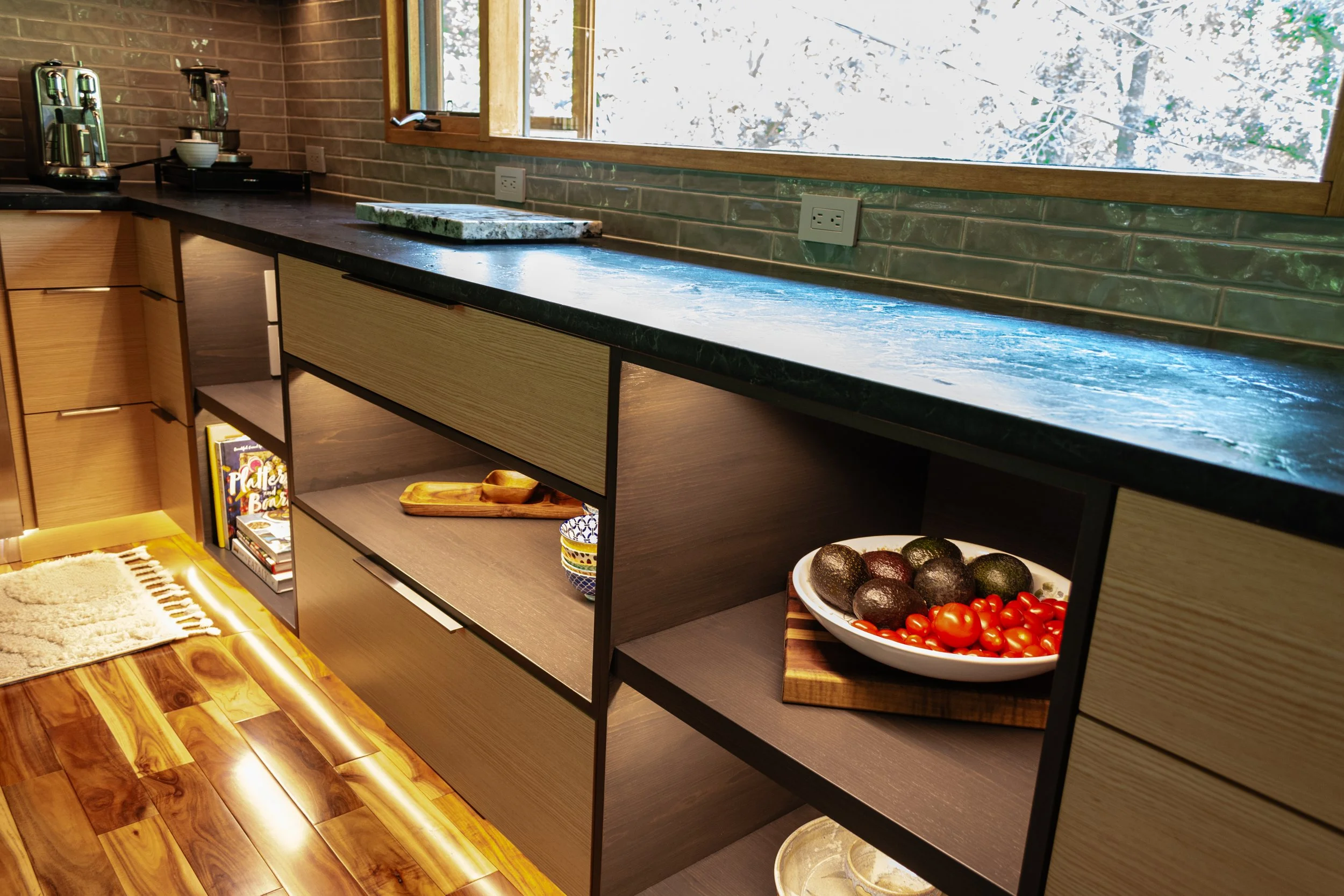 Modern kitchen with wooden cabinets, a green marble countertop, and a window showing trees outside. The shelves contain kitchenware and books.