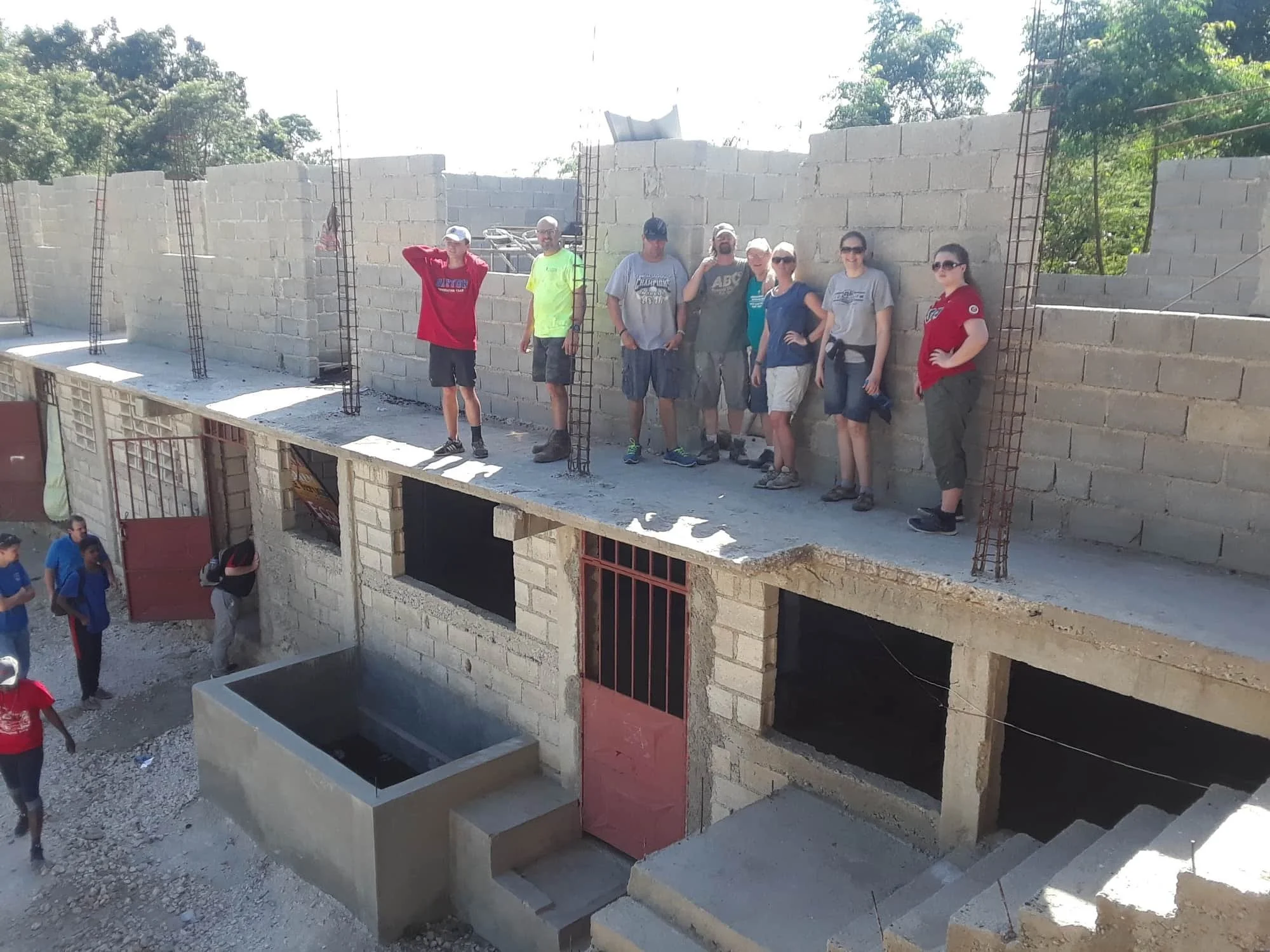 A group of people standing on an upper level of a building under construction, with concrete blocks and rebar visible, overlooking a lower area with people walking.