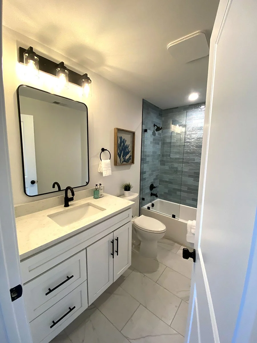 Modern bathroom with white vanity, black fixtures, rectangular mirror, framed artwork, dual faucet, soap dispenser, potted plant, toilet, and bathtub with glass enclosure and gray tile walls.