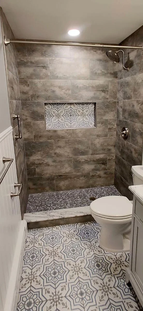 Bathroom with a walk-in shower featuring grey tiles, a patterned accent tile, and a clear glass mosaic bathroom floor. There is a supply of towels on a rack on the left wall, a toilet on the right, and a small vanity with a marble top.