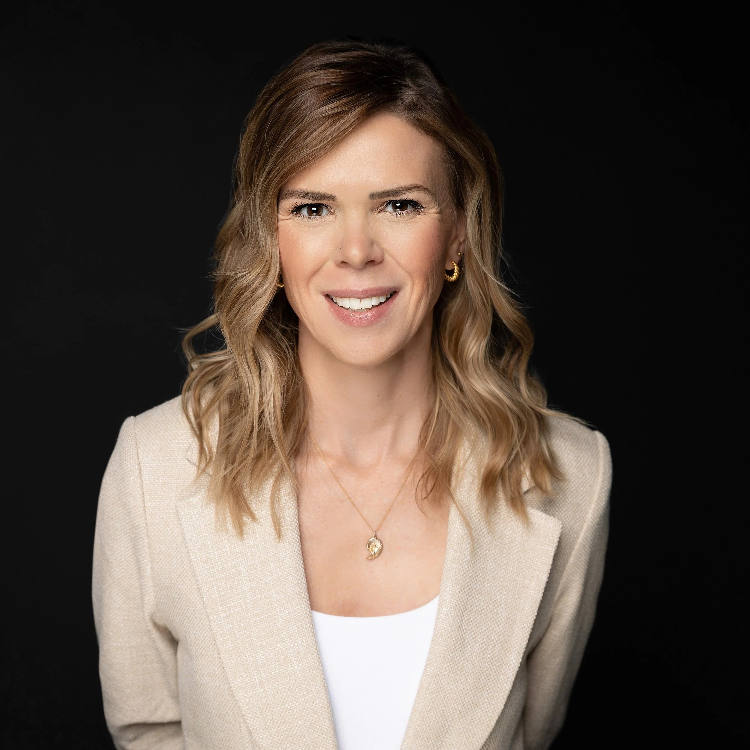 Headshot of a woman with shoulder-length wavy blonde hair, smiling, wearing a beige blazer, gold jewelry, and a white top against a black background.