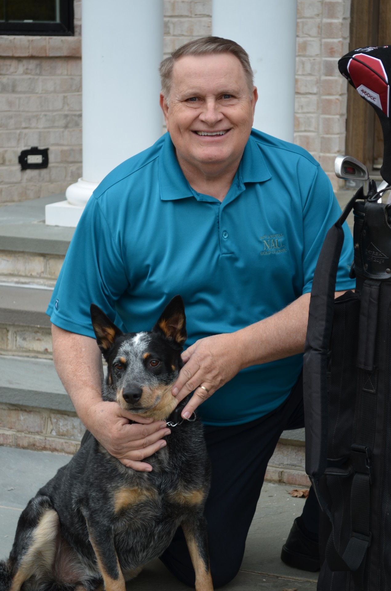 Michael Sampson in a blue polo shirt kneeling next to a black and tan Australian cattle dog, holding its collar. They are outside in front of a brick house with white columns.
