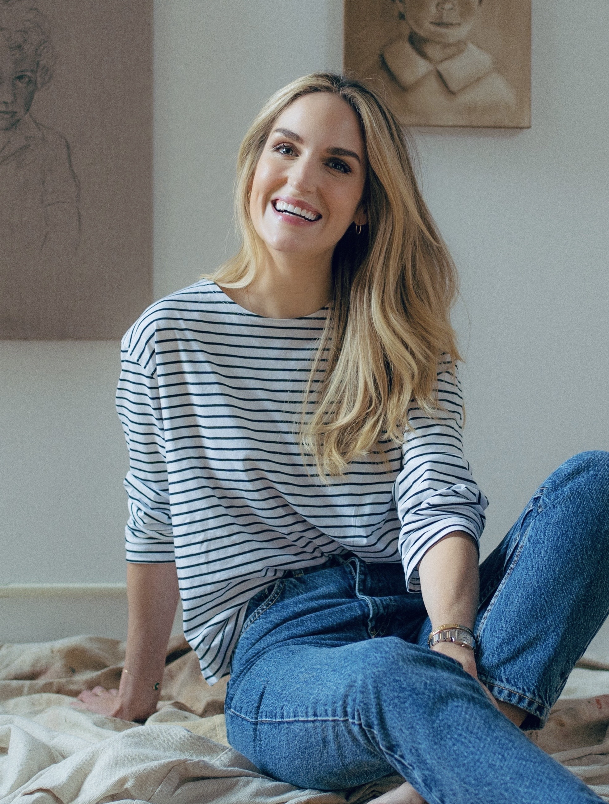 Lauren Phelps, a young woman with long blonde hair smiling while sitting on a bed in a casual striped shirt and jeans, with artwork hanging on the wall behind her.