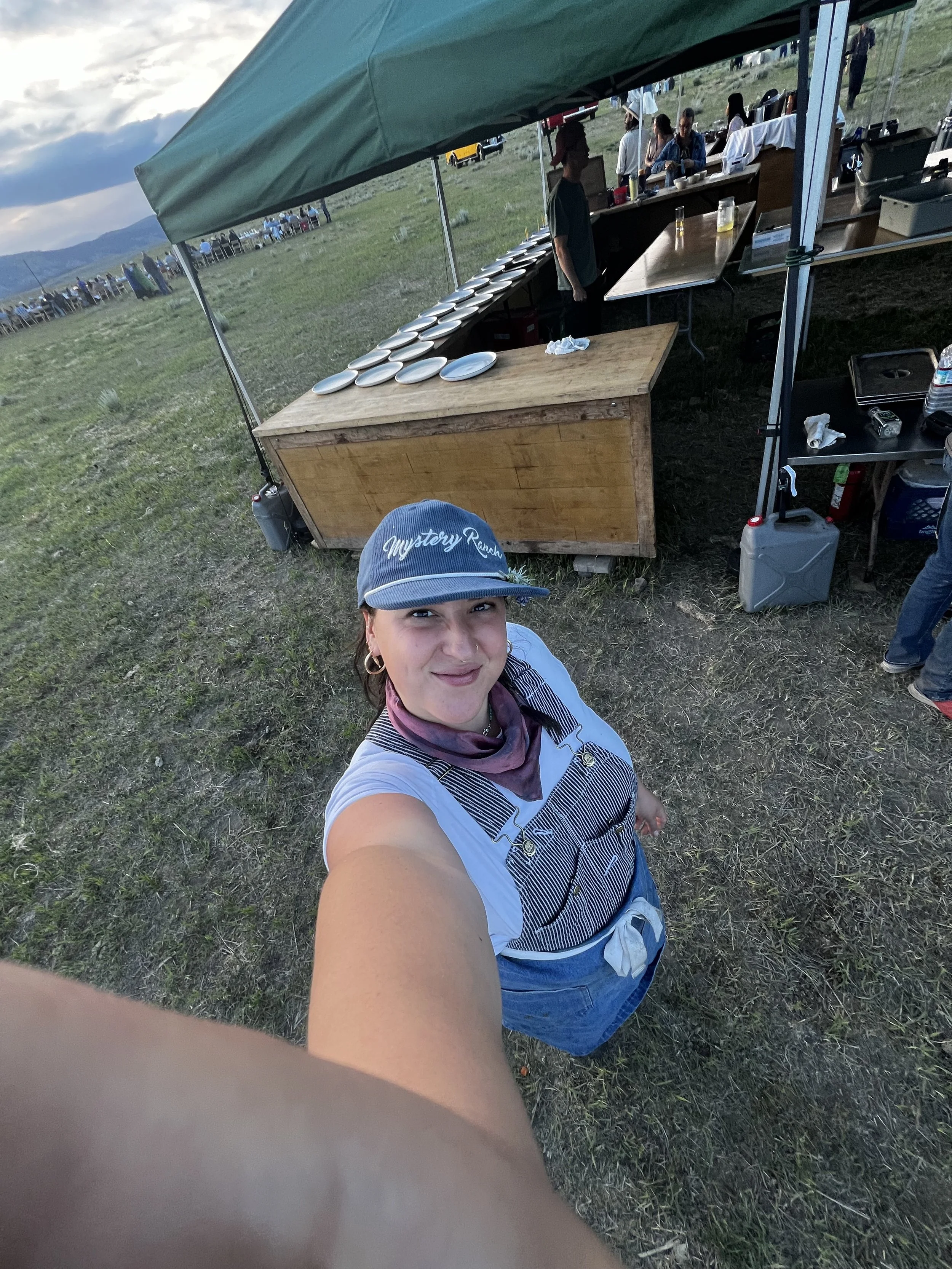 A woman wearing a blue cap and striped apron taking a selfie at an outdoor event with a food stand behind her, set in a grassy field.