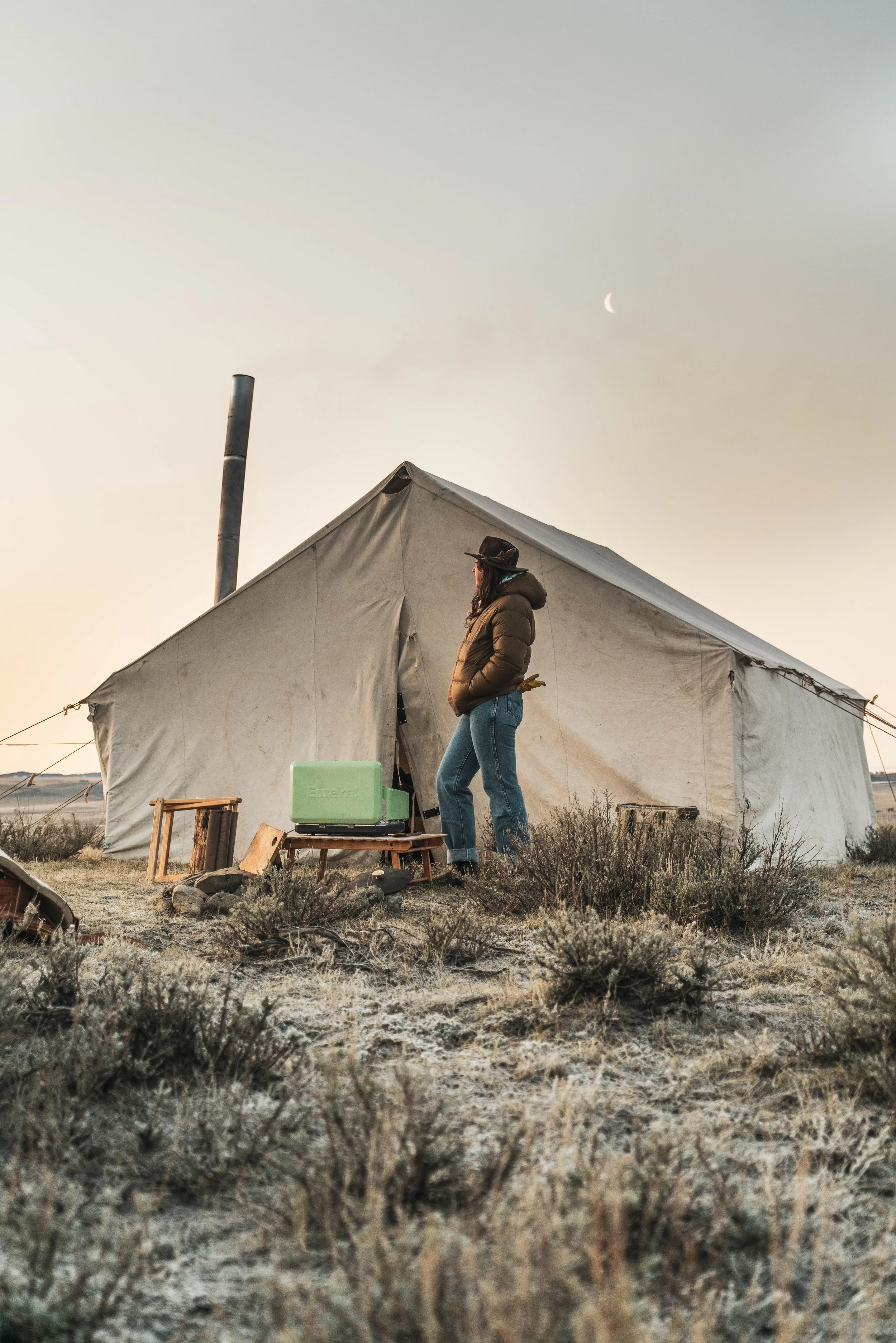 A woman standing outside a large canvas tent in a desert landscape during sunset or sunrise, with a crescent moon visible in the sky, wearing a jacket, jeans, a fedora, and glasses.