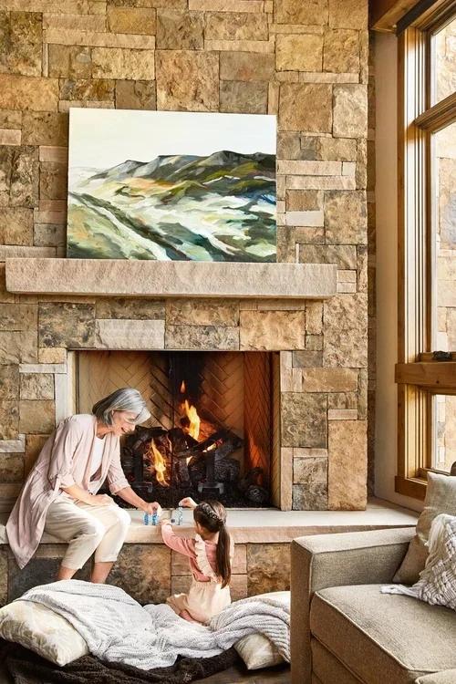 An elderly woman and a young girl sitting by a fireplace in a cozy living room, with a landscape painting above the fireplace and a large window to the right.