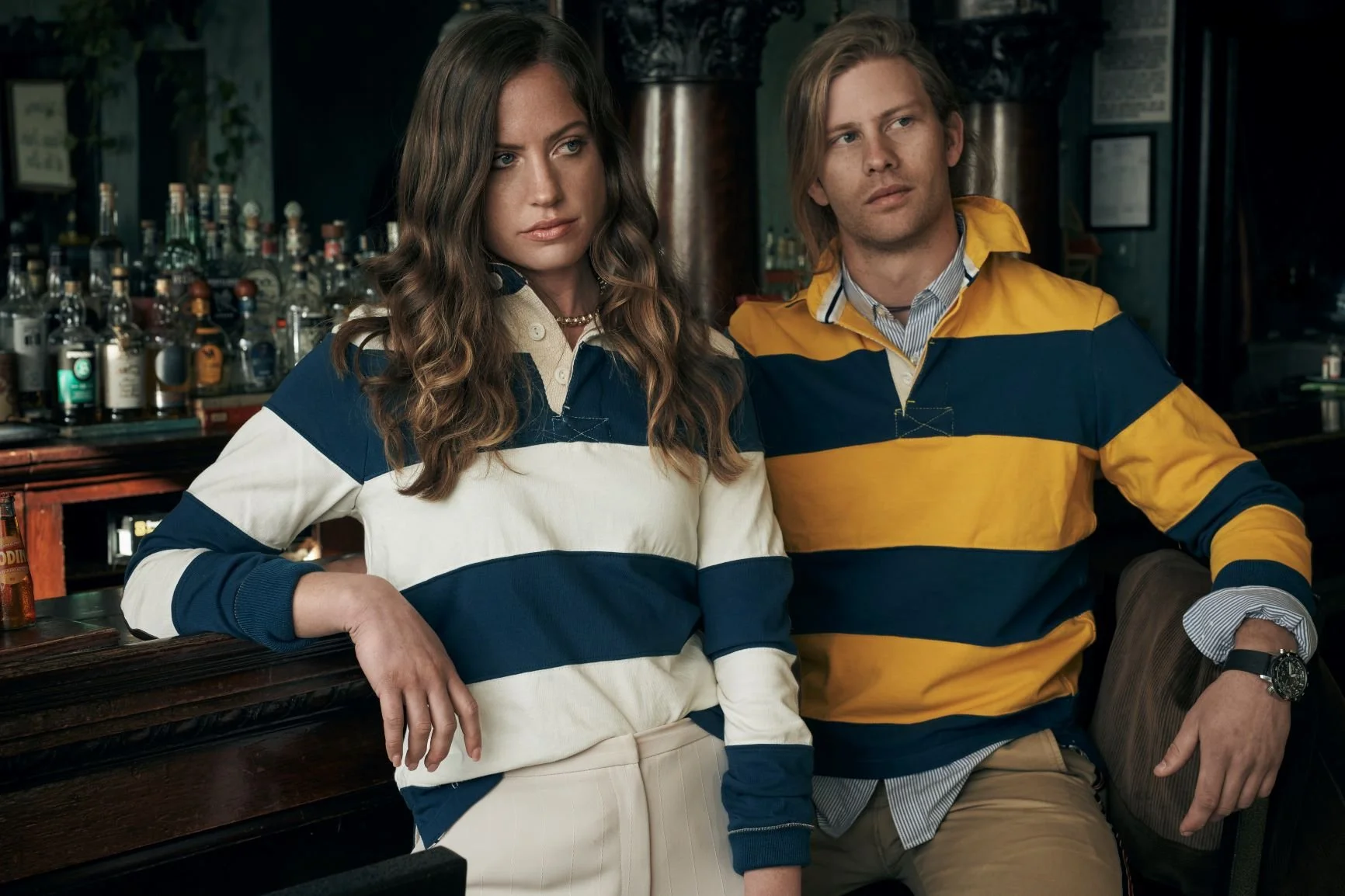 Two young adults sitting at a bar, both wearing striped Maroon Bells rugby shirts, with bottles and glasses in the background.