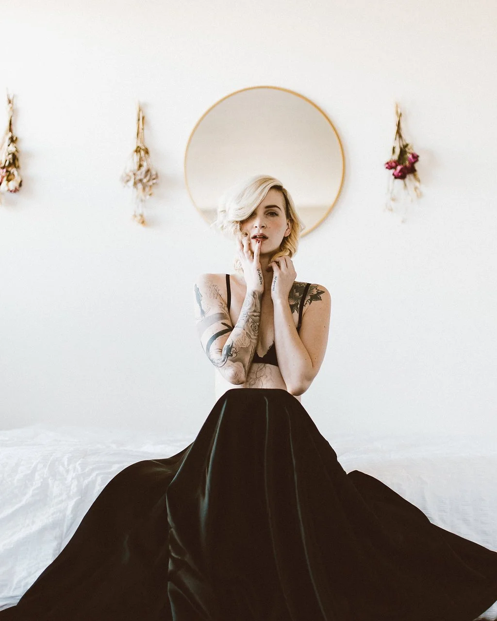 A woman with short blonde hair and tattoos on her arms and chest, wearing a black top and a black skirt, sitting on a white bed in a minimalistic room with a round mirror and dried flowers on the wall behind her.