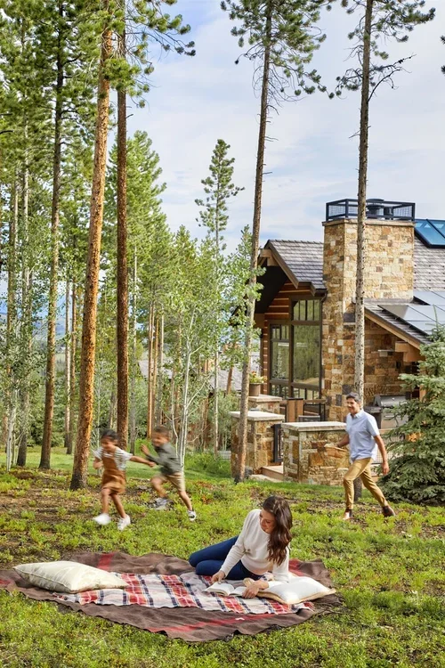 A woman sitting on a blanket reading a book with children playing chase in a wooded yard near a stone house.