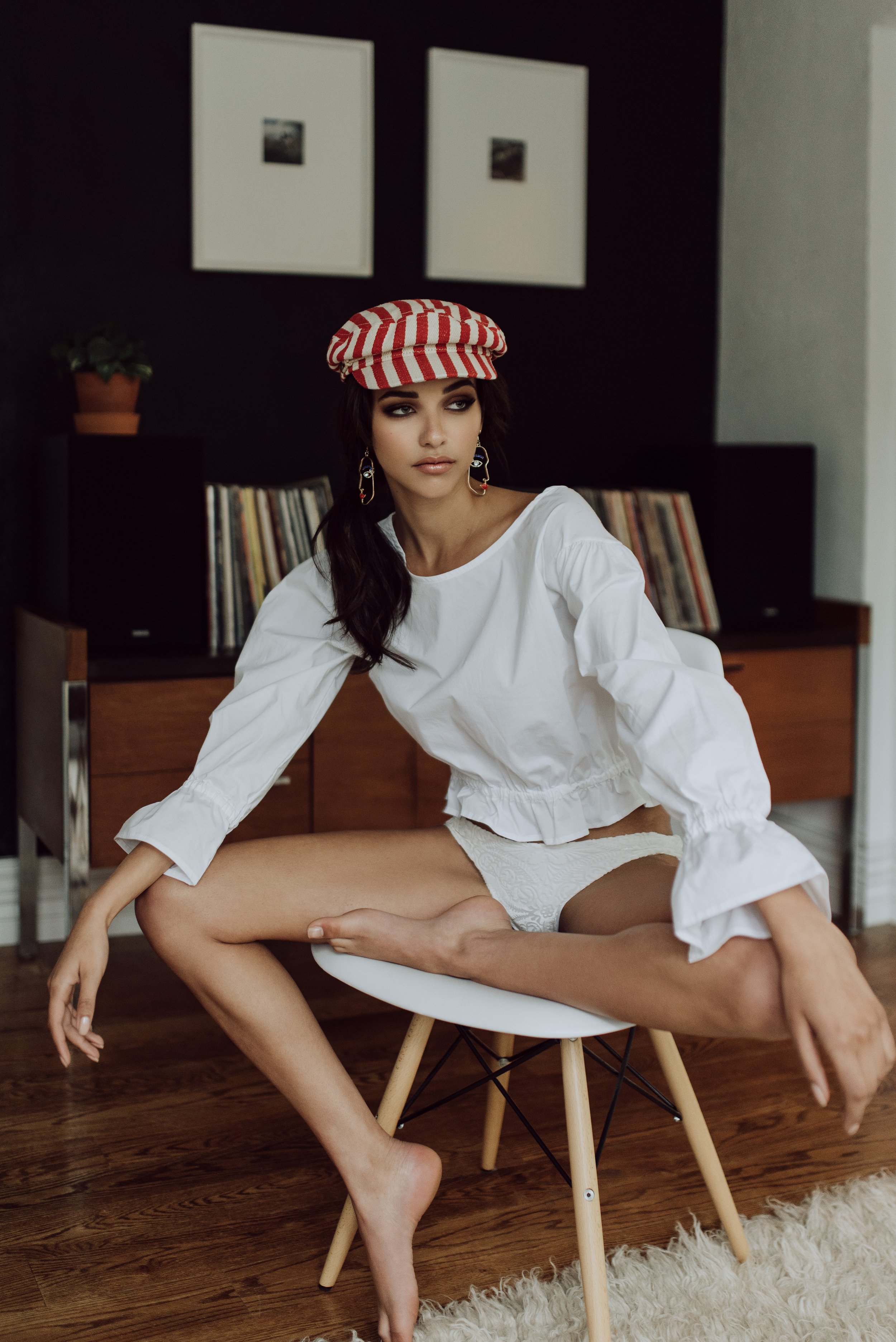 A woman with dark hair, wearing a striped red and white cap, large earrings, a white blouse, and white underwear, sitting on a modern stool in a room with a wooden floor, a black and white wall, and framed pictures and plants in the background.