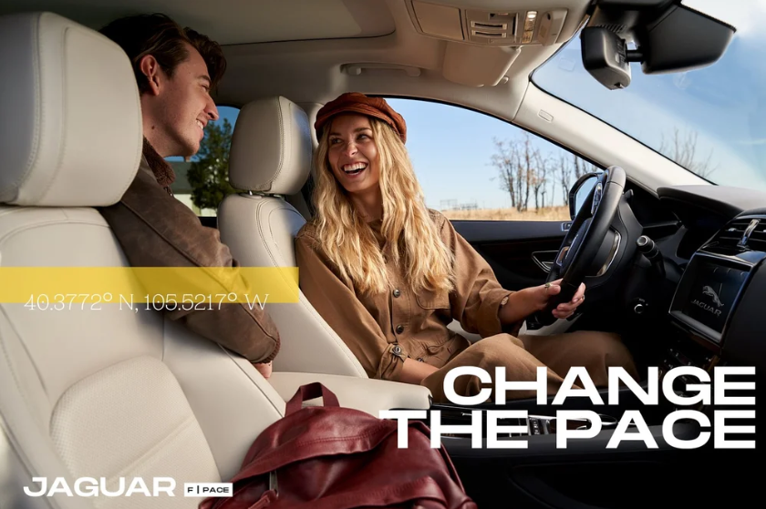 Two people in a car, with the woman driving and smiling at the man in the passenger seat during daytime, with trees visible outside. Text overlay says 'Change the Pace' and 'Jaguar 1 Pace'.