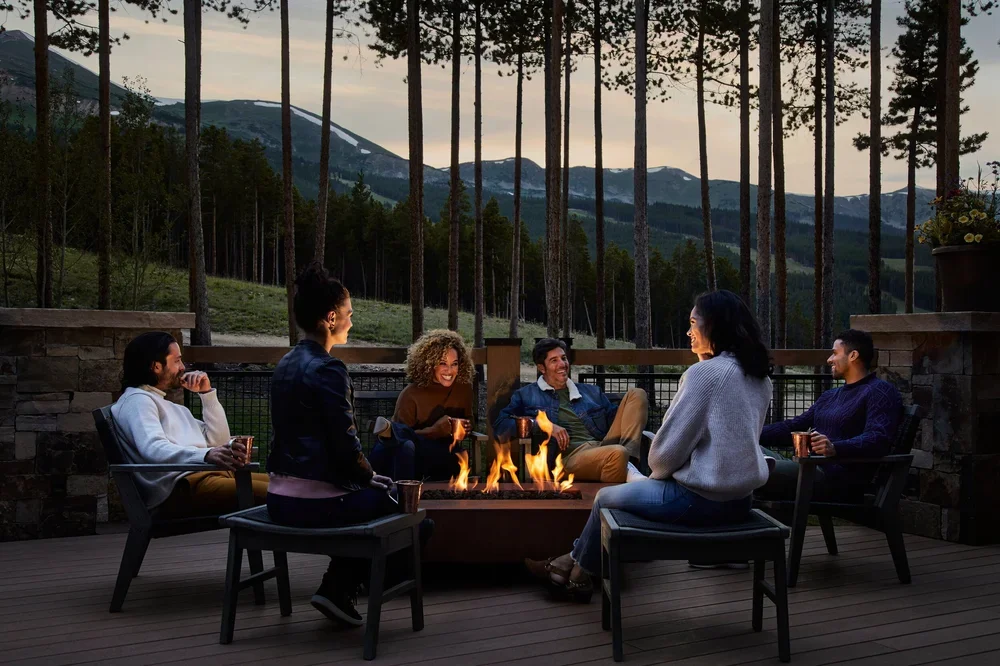 A group of six friends sitting around a firepit on a wooden deck, enjoying drinks, with a mountain landscape and tall trees in the background during dusk.