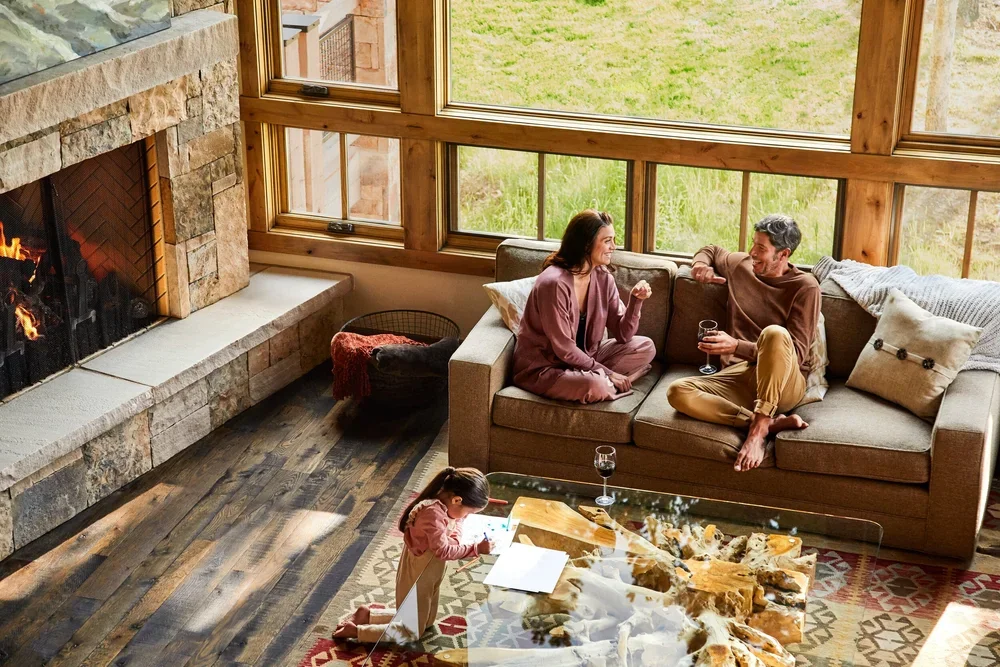 A family of three enjoying quality time in a cozy living room near a fireplace with a roaring fire. A woman and a man are sitting on a sofa, talking and drinking wine, while a young girl is on the floor drawing with a crayon on paper.