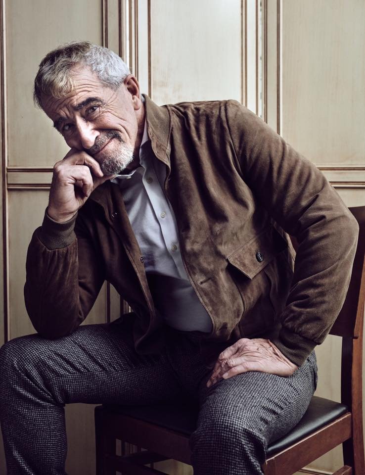A senior man with gray hair and a beard sitting on a wooden chair, resting his right elbow on his knee and his chin on his hand, smiling and looking at the camera. He is wearing a brown suede jacket over a light-colored shirt and patterned pants, with a wooden-paneled wall in the background.