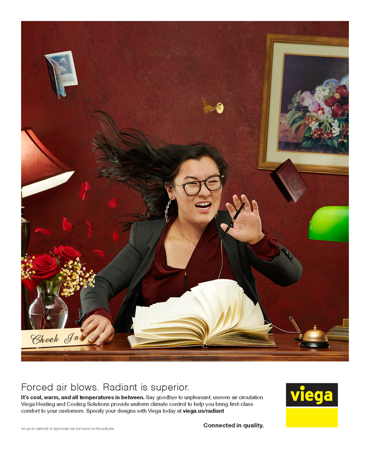 A woman with long dark hair, glasses, and earrings sits at a desk with an open book in front of her, hair flying, eyes closed, and expression of discomfort. The background features a red wall with a framed picture of flowers, a green desk lamp, a red wall lamp, and falling rose petals. There are flowers in a vase on the desk, and a sign that reads 'Check In'.