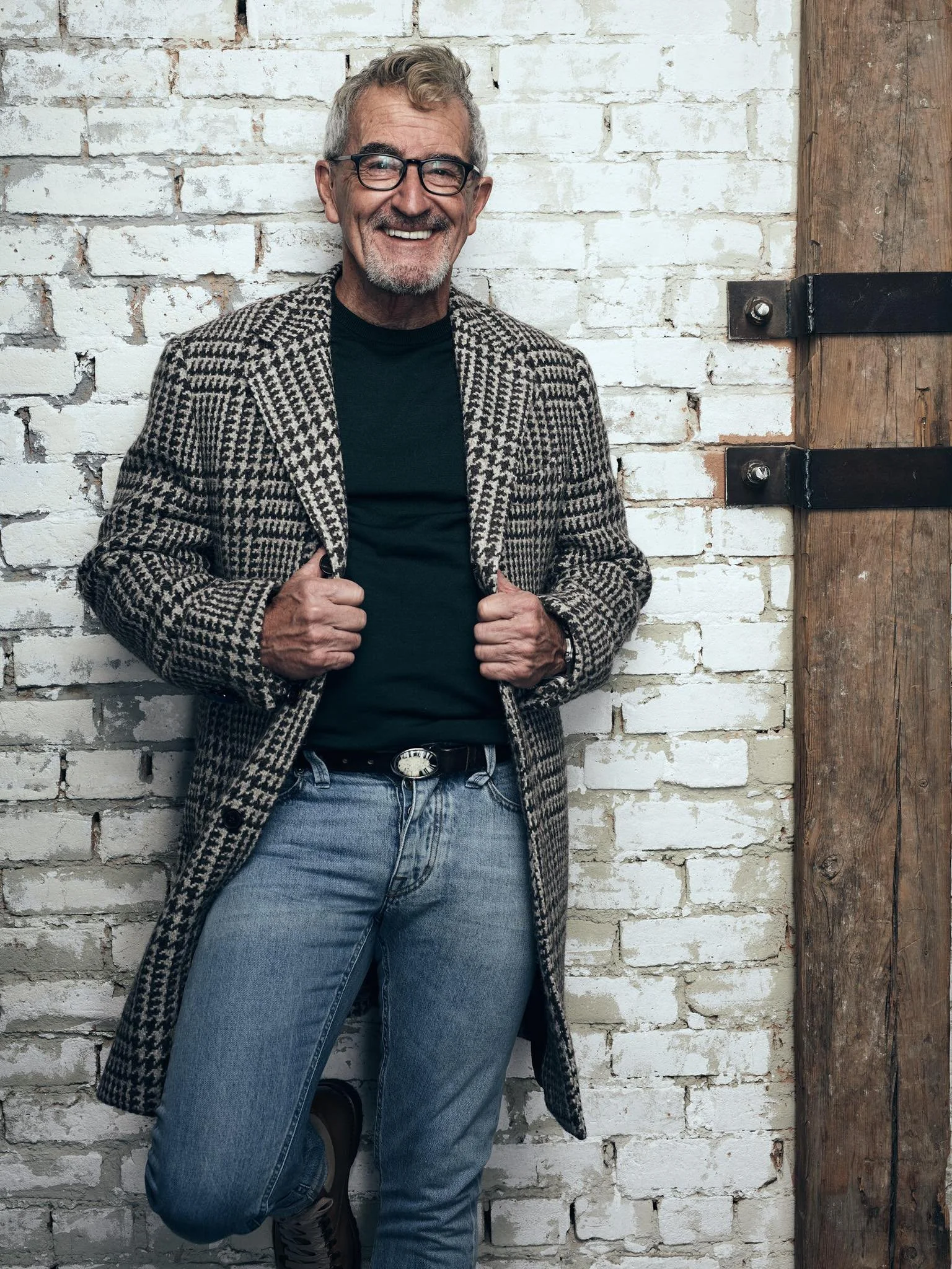 An older man with gray hair, glasses, and a beard smiling, standing against a white brick wall, wearing a houndstooth patterned coat, black shirt, jeans, and a belt.